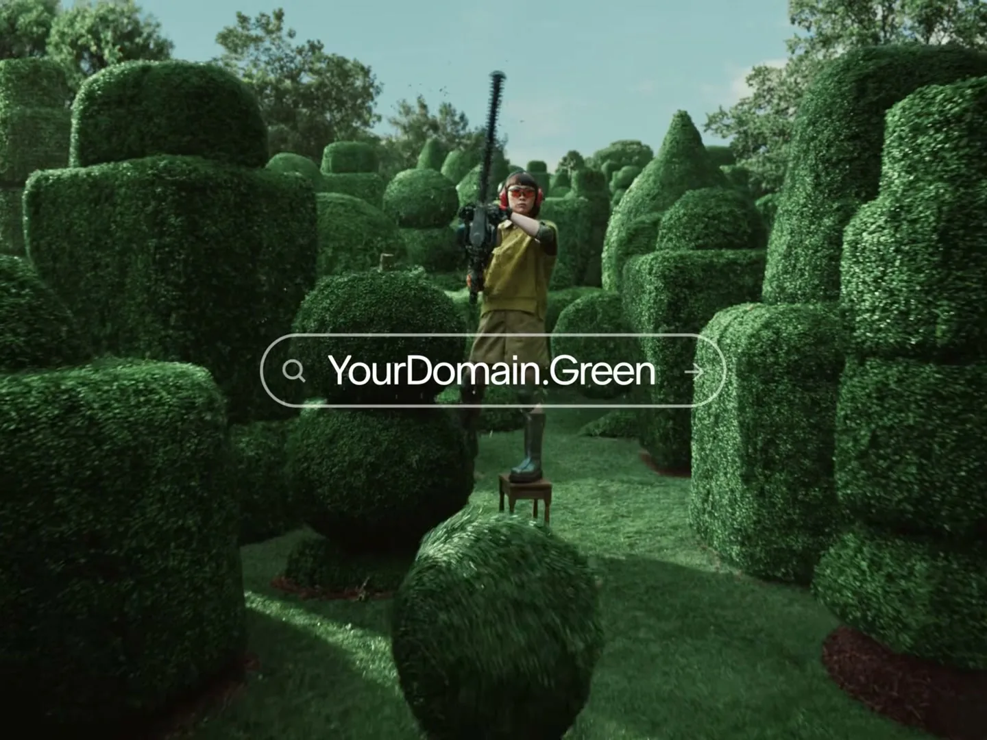 A person in a green outfit and ear protection stands on a stool, trimming a large, round topiary with a hedge trimmer in a whimsical garden.
