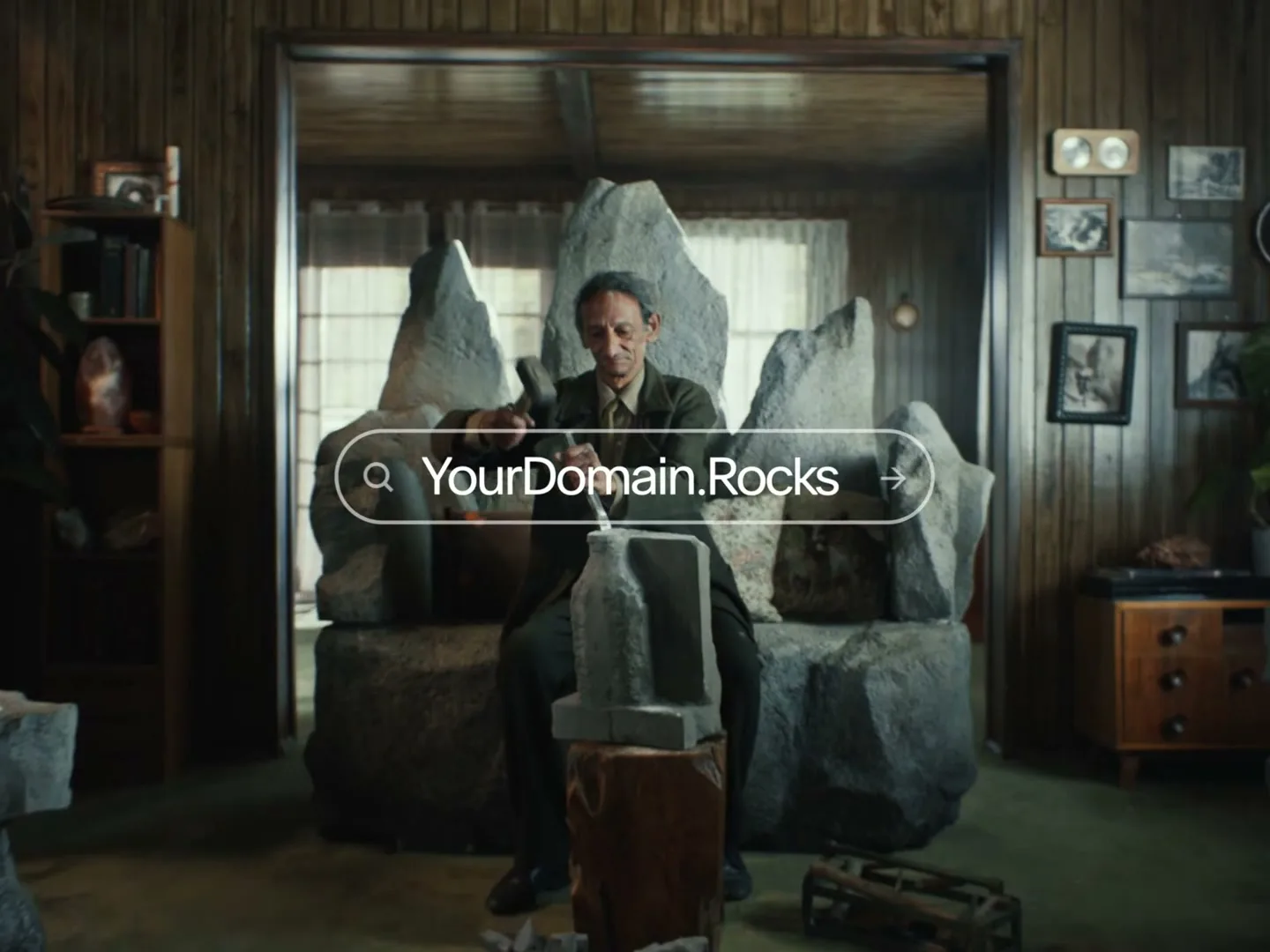 An older man with dark skin and a dark suit sits on a stone throne, carving a small stone sculpture in a rustic room.