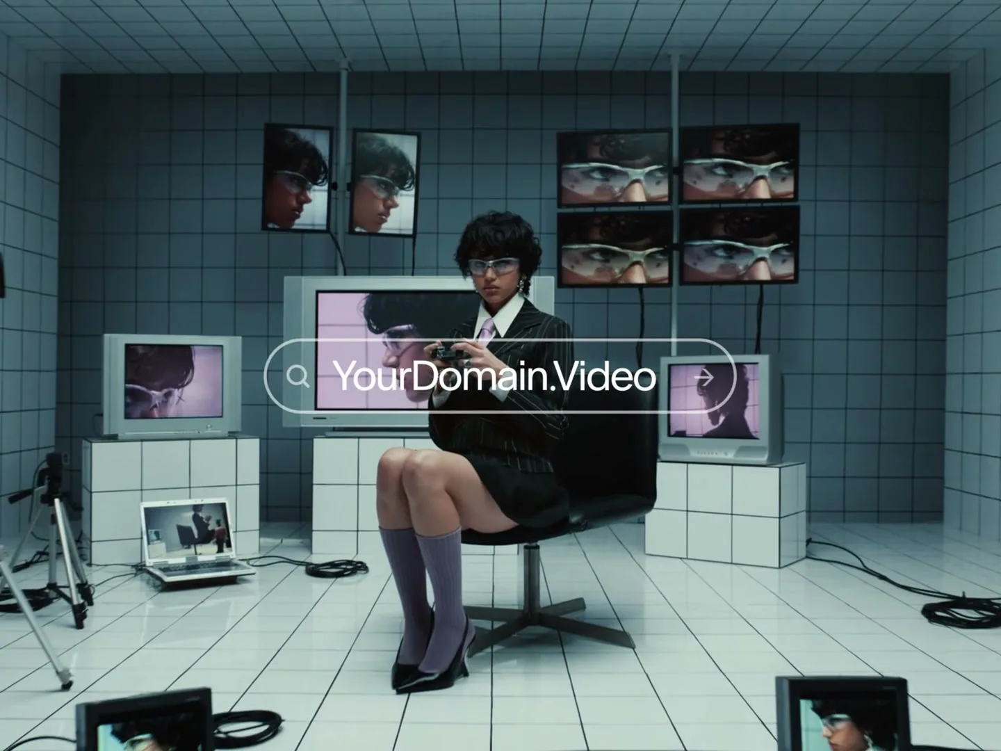 A young woman in a suit and glasses sits in a room surrounded by monitors displaying her image, holding a video game controller, with a search bar overlayed on screen.