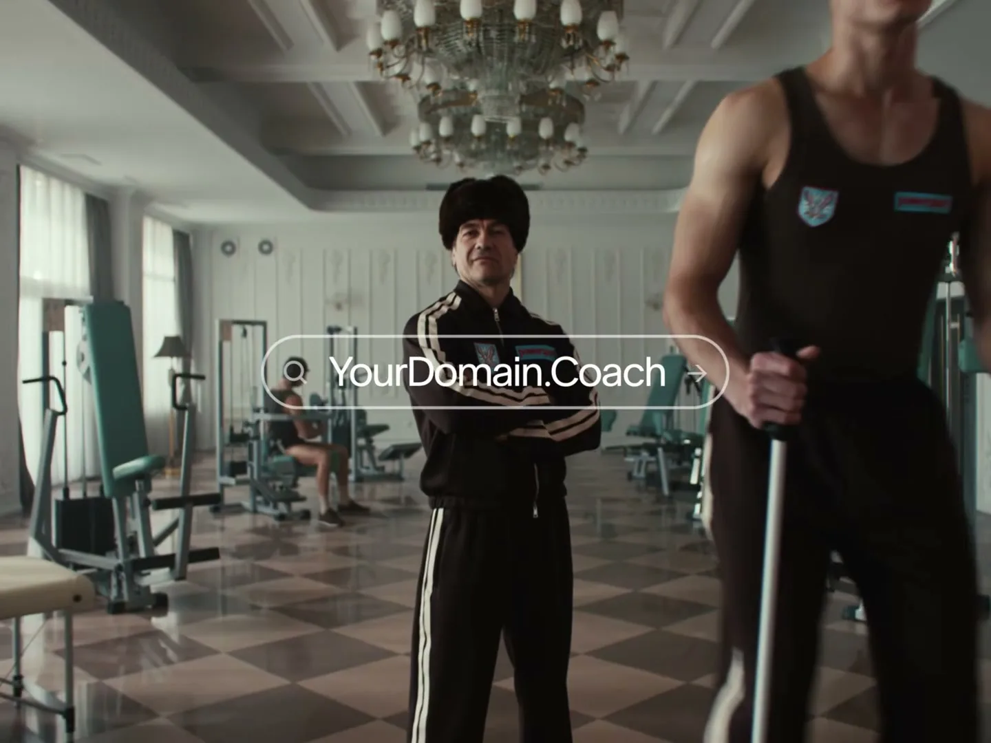 A man in a fur hat and tracksuit stands with arms crossed in a gym, as a younger man walks past him on a ski machine, with a domain search bar overlayed.