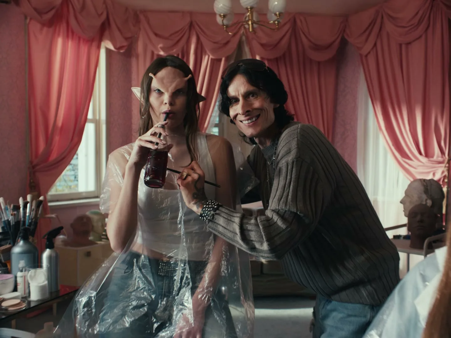A woman with prosthetics and a man with long hair smile at the camera while the woman drinks from a straw in a pink room.