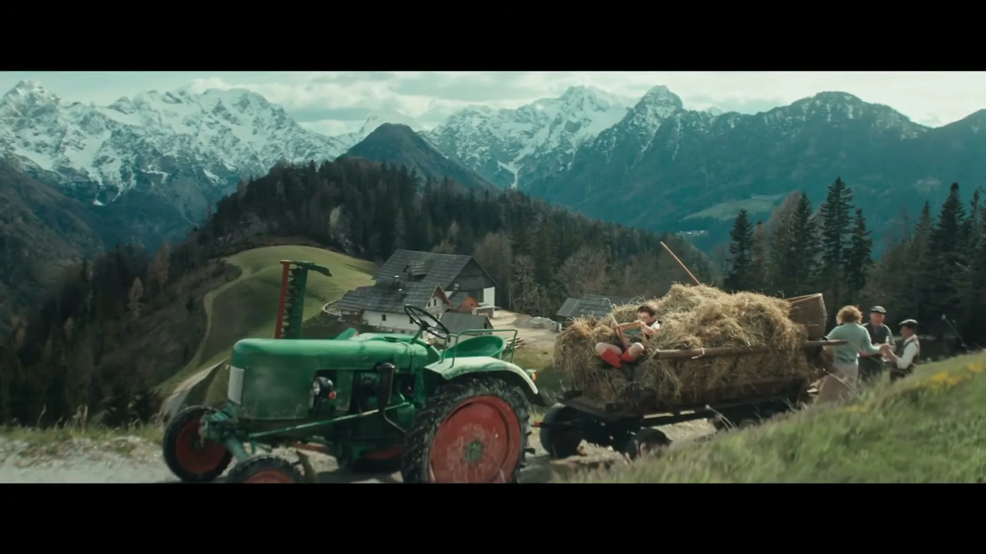 A green tractor pulls a hay-filled cart with a child and two men on a winding road, set against a backdrop of snow-capped mountains and a village.