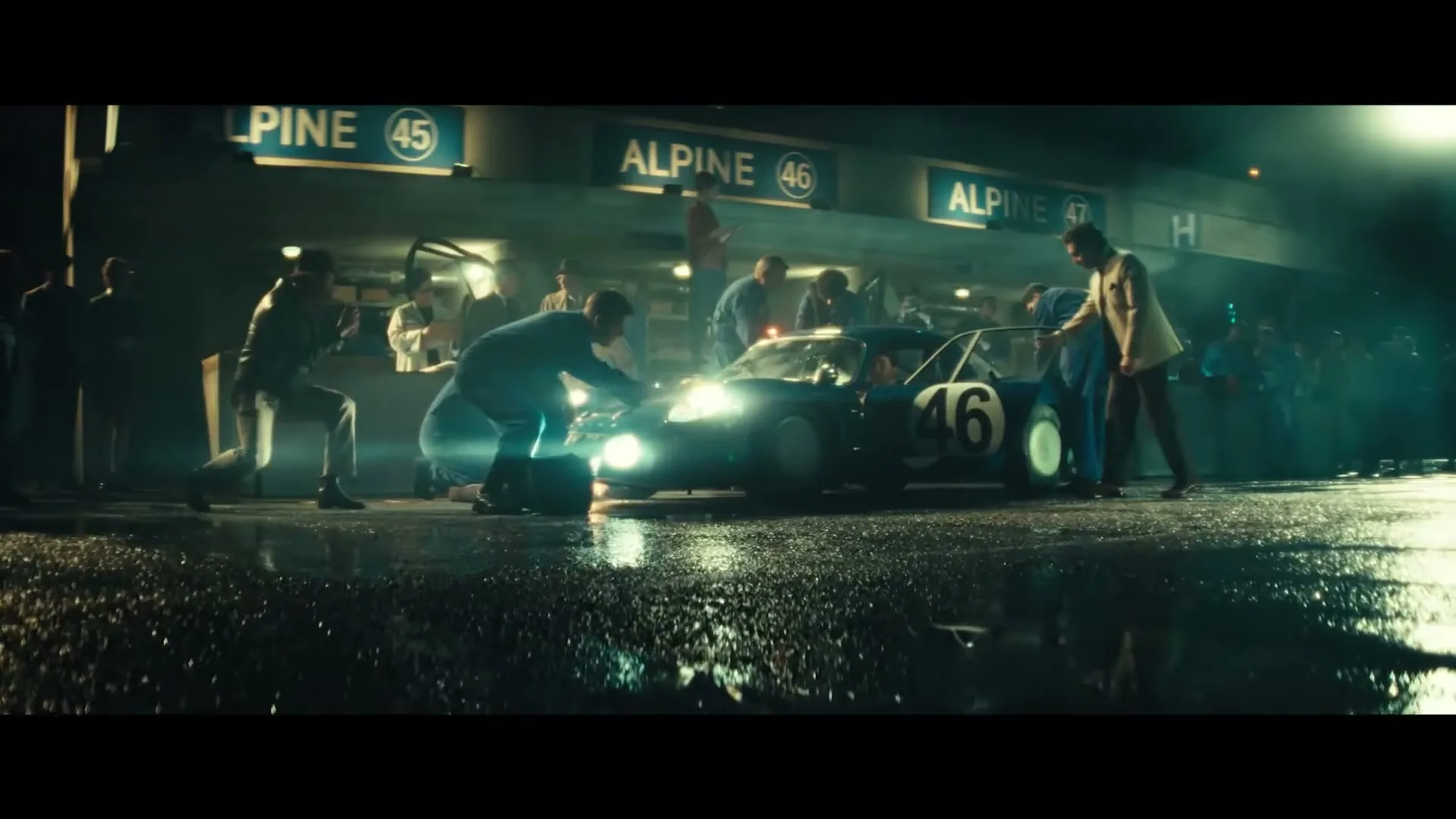 A vintage blue race car with number 46 is surrounded by a pit crew and spectators at night, with bright headlights illuminating the wet ground.