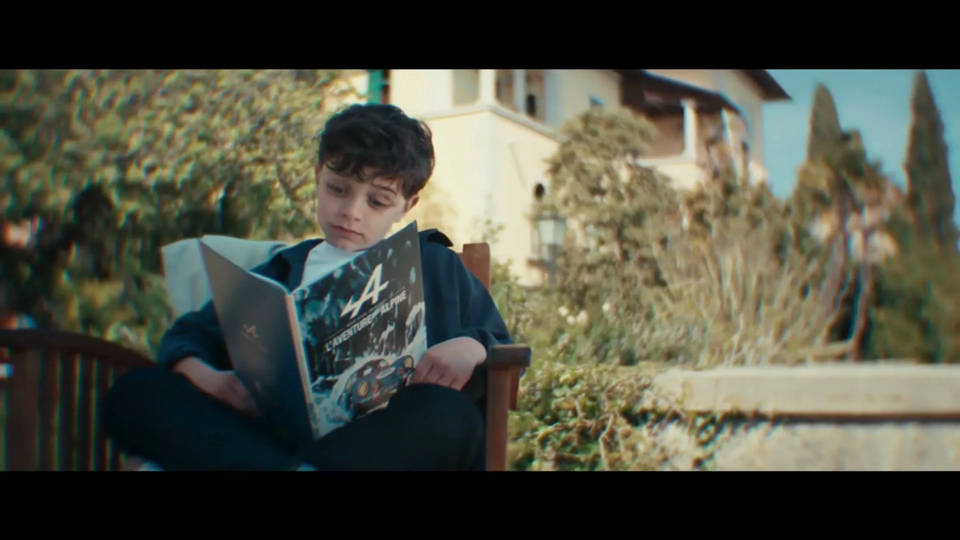 A young boy with dark curly hair sits cross-legged in a wooden chair outdoors, reading a book with a car on the cover, looking up thoughtfully.