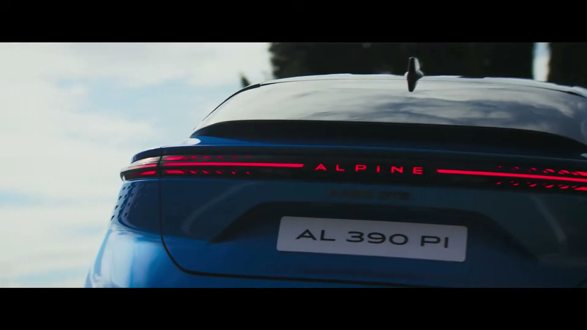 The rear of a blue Alpine car with illuminated taillights and license plate is shown from a low angle, moving slightly on a sunny day.