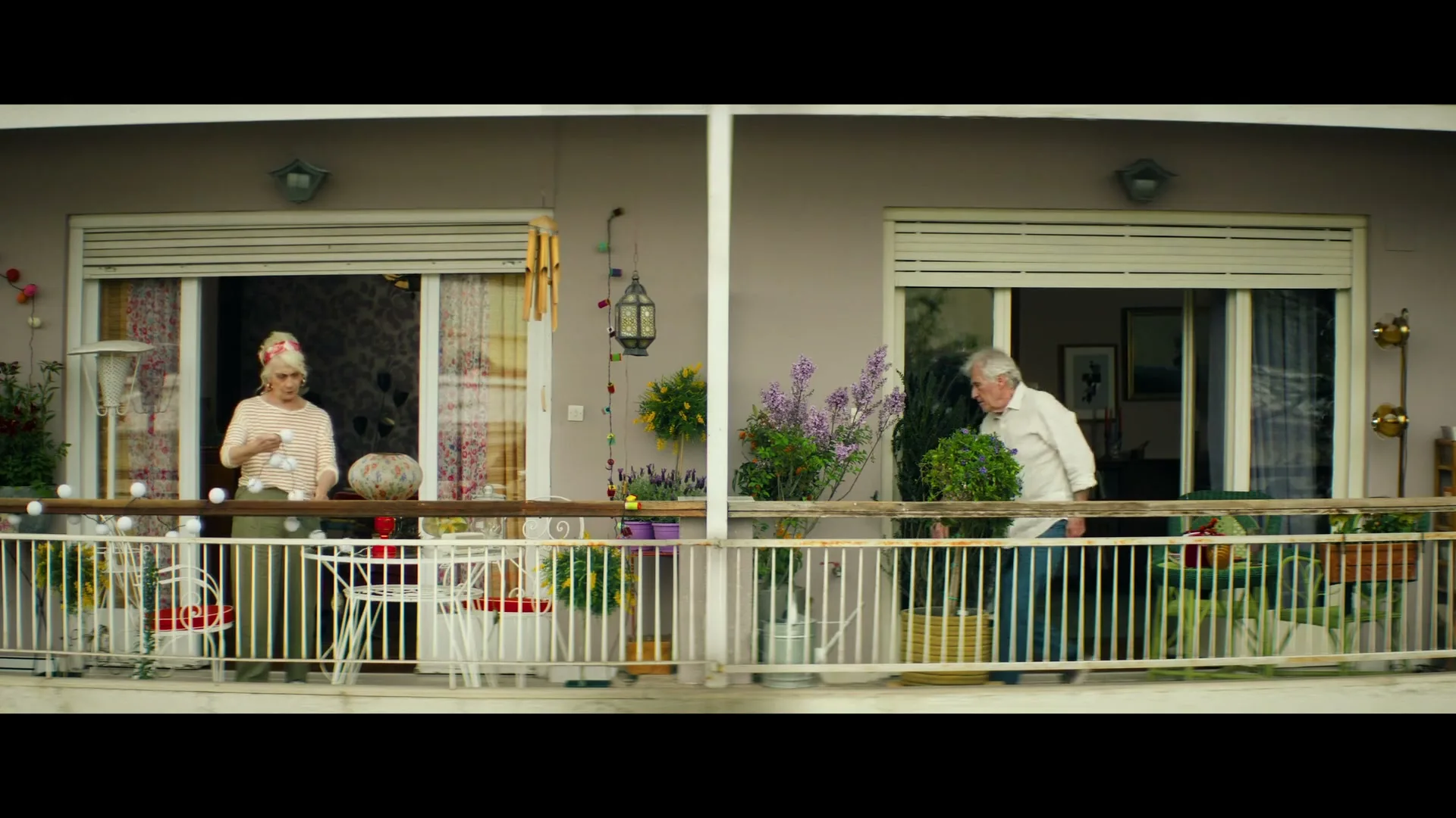 An elderly woman in a striped shirt and a man in a white shirt are on separate balconies, tending to plants and looking at each other.