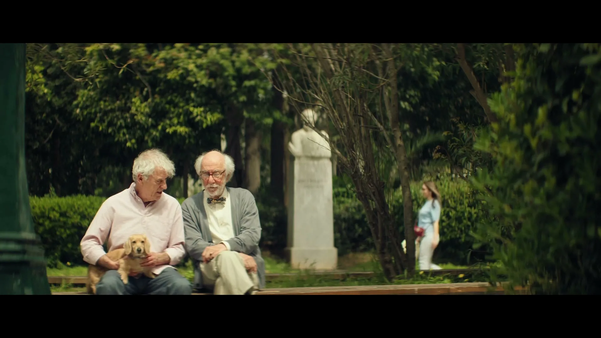 Two elderly men sit on a park bench, one holding a small dog, as a young woman walks by in the background.