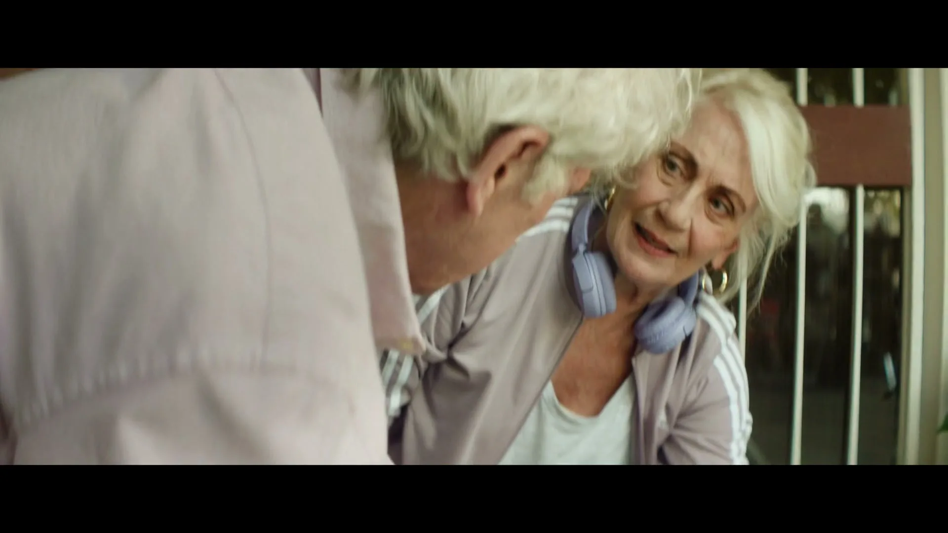 An older man and woman are on the floor, picking up groceries that have spilled from a brown paper bag, with a pineapple and tomatoes visible.
