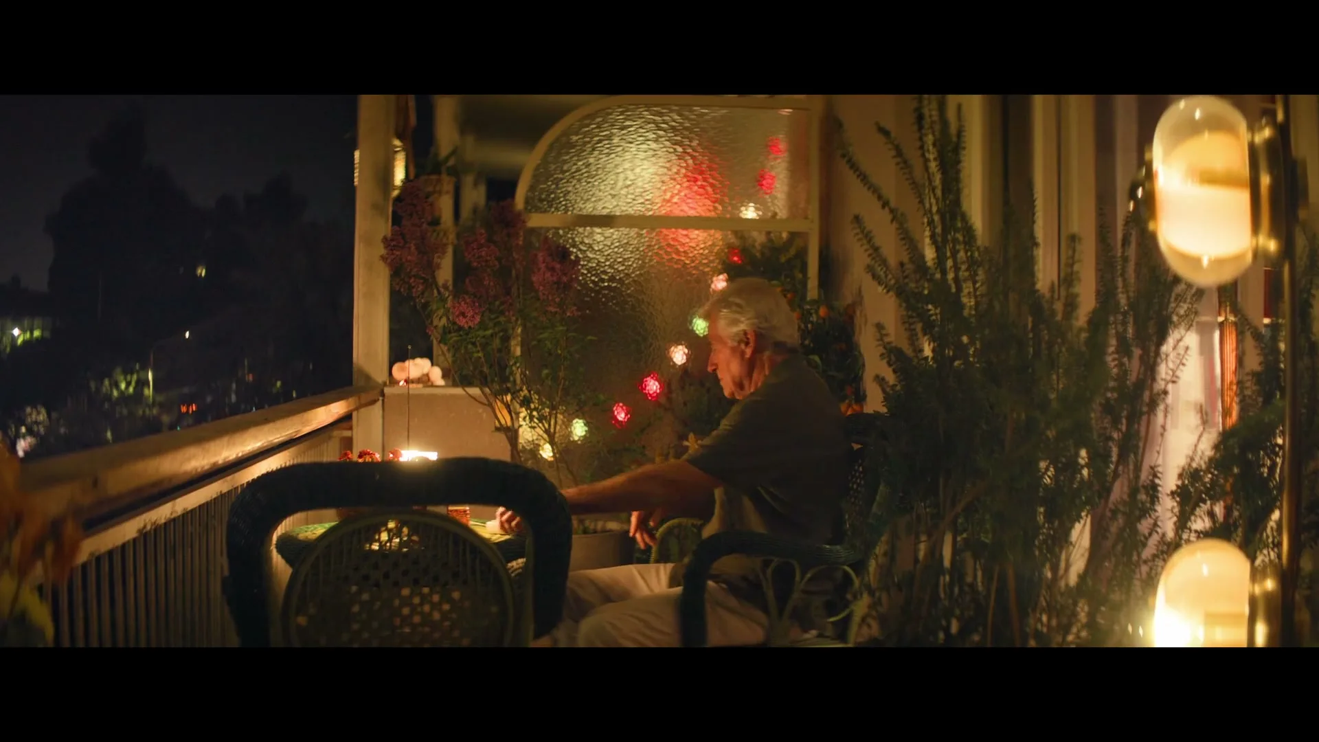 An elderly man with white hair sits on a balcony at night, surrounded by plants and string lights, looking out into the darkness.
