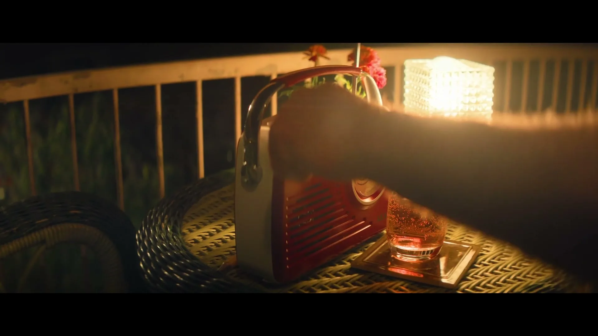 A man's hand adjusts a vintage red radio on a wicker table next to a drink and a lamp on a balcony at night.