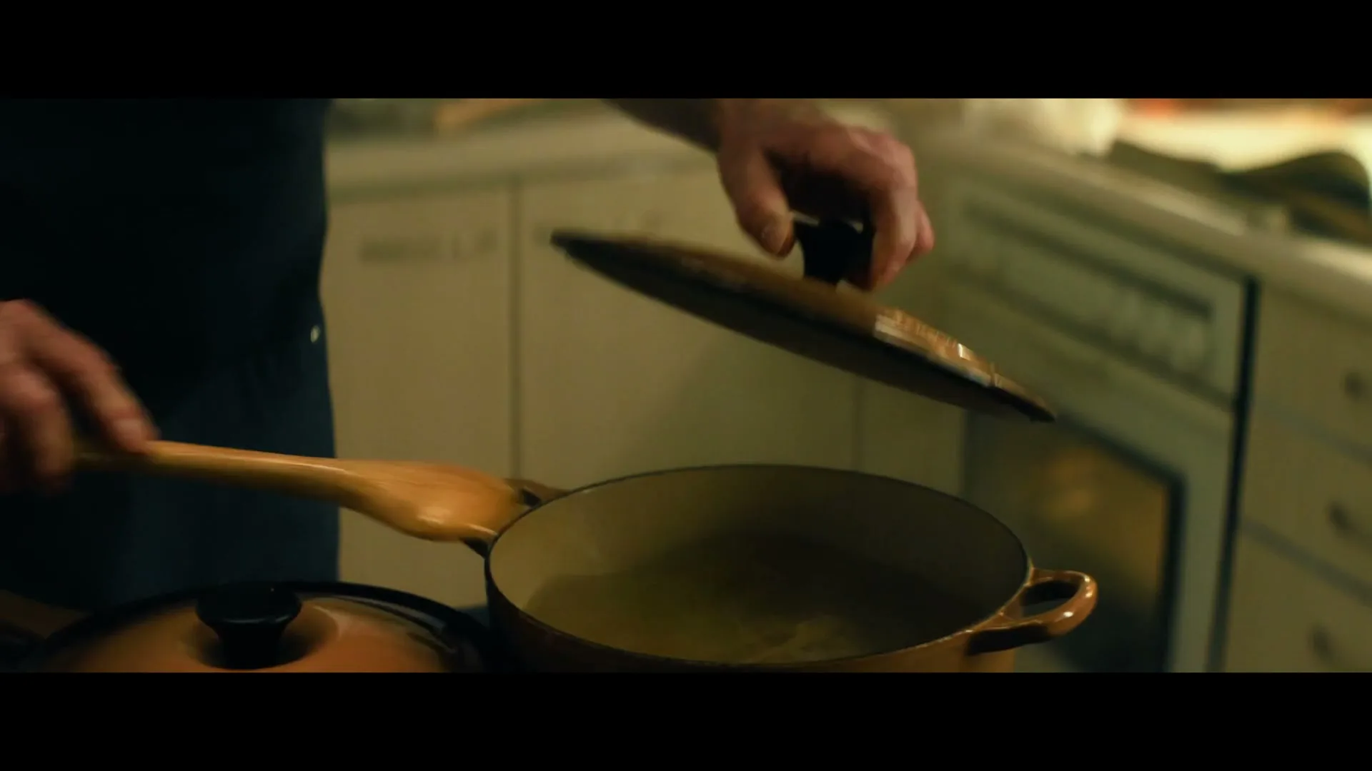 A man's hands are seen stirring pasta in a pot with a wooden spoon, then placing a lid on another pot in a kitchen.