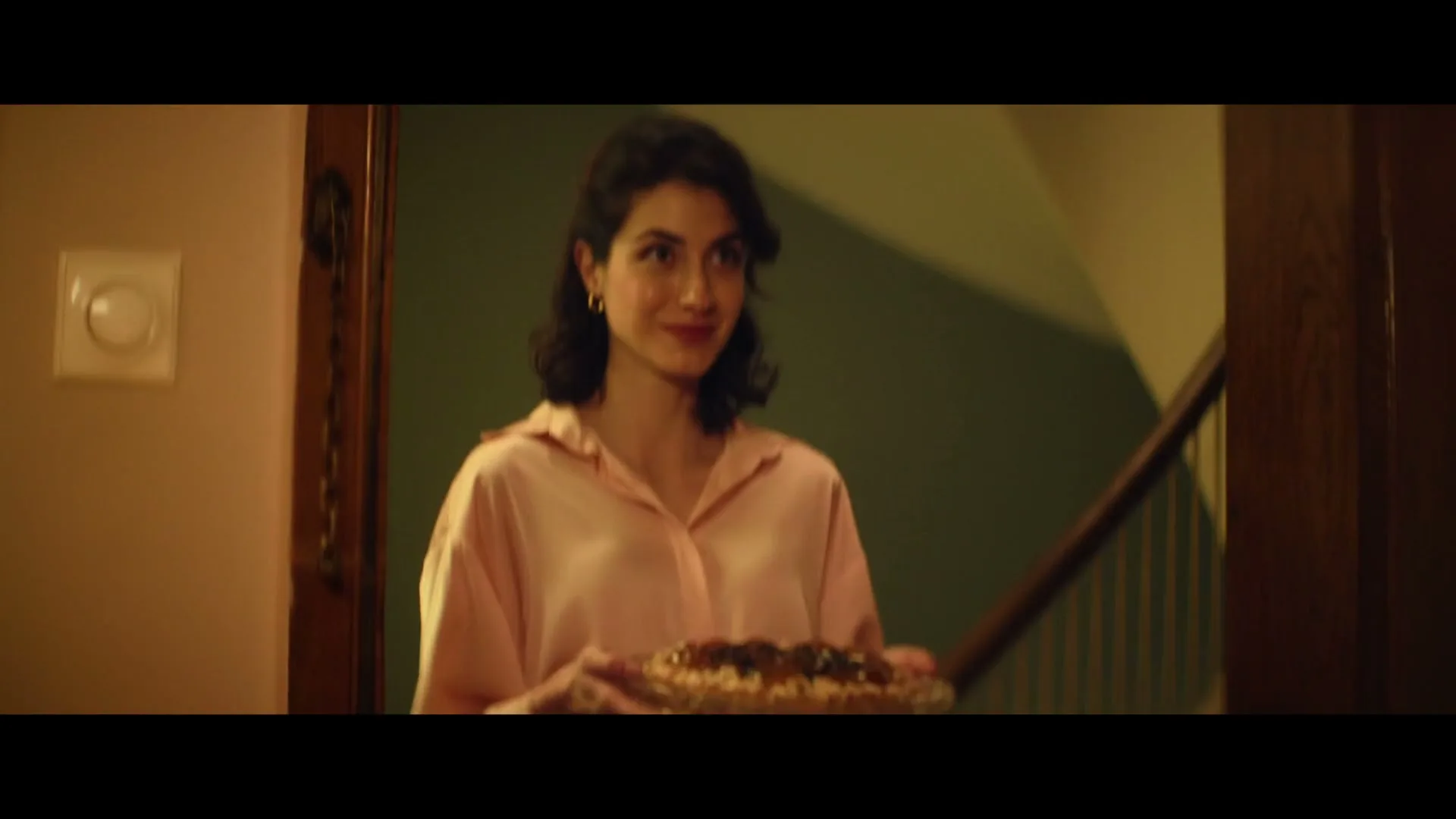 A woman in a pink shirt opens a wooden door, revealing herself holding a pie, with a staircase visible in the background.