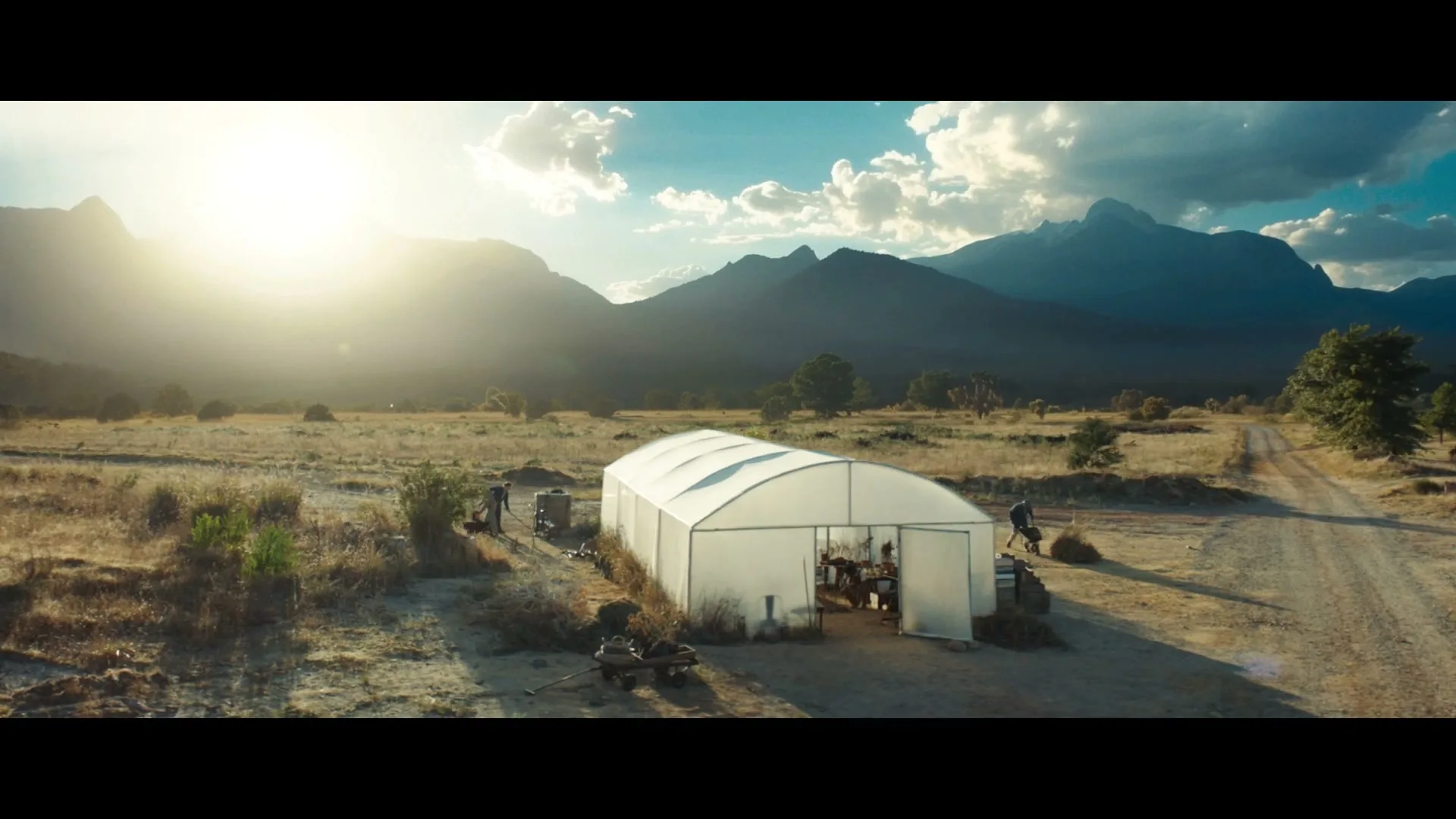 A wide shot of a white greenhouse in a dry, rural landscape with mountains in the background under a bright sun.
