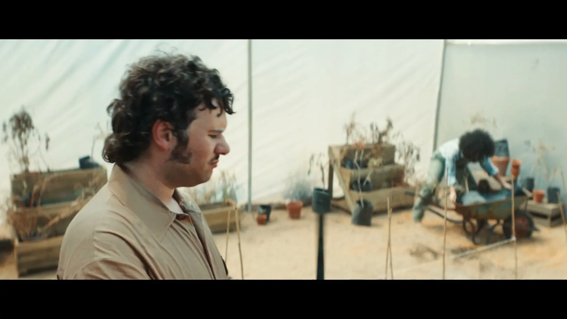 A man with curly hair and a mustache looks down sadly, holding a stick in a greenhouse with dead plants, while another person works in the background.