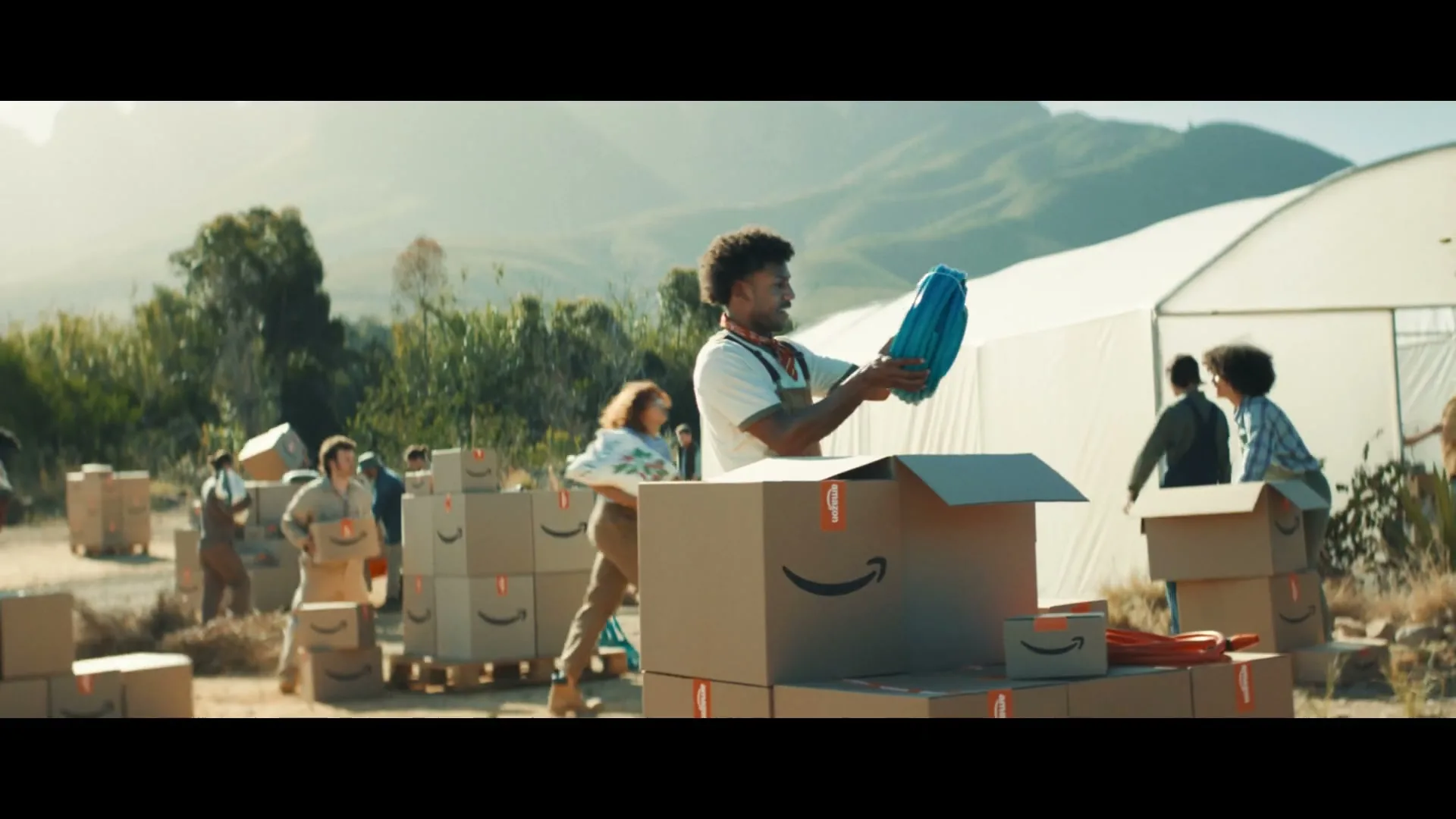A man in an apron holds up a blue hose while packing Amazon boxes outdoors, surrounded by other workers and boxes near a greenhouse.
