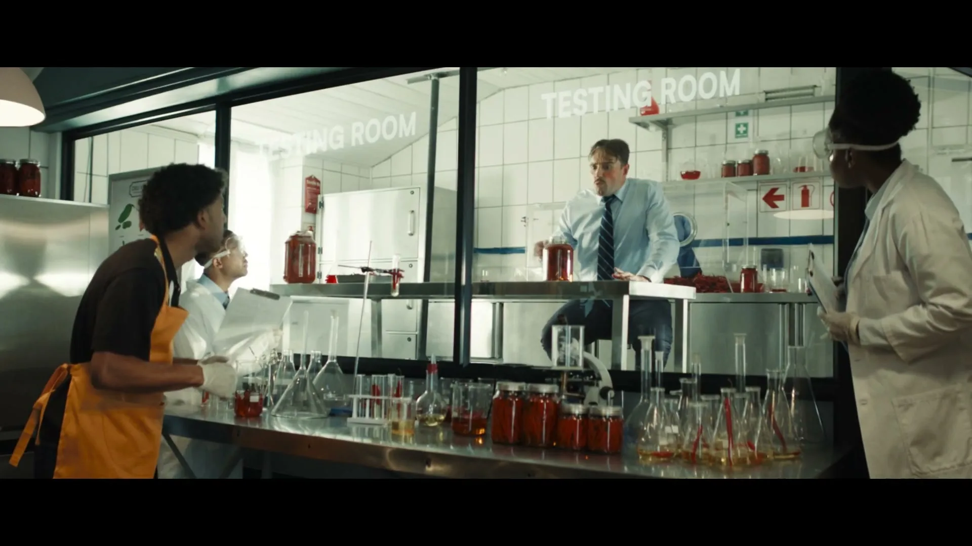 A man in a shirt and tie sits on a counter in a testing room, yelling at two scientists and a man in an apron in a lab.