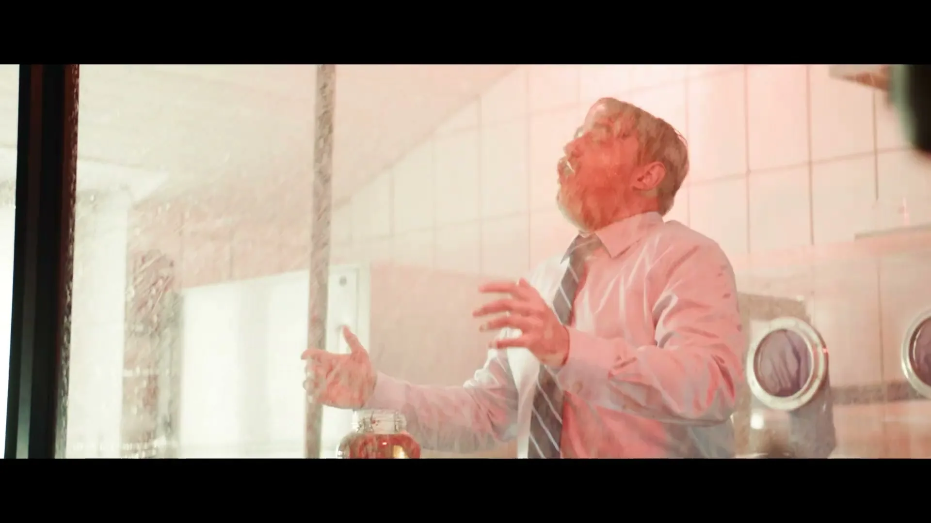 A man in a light blue shirt and striped tie is being sprayed with liquid in a sterile-looking room, holding a jar of pickles.