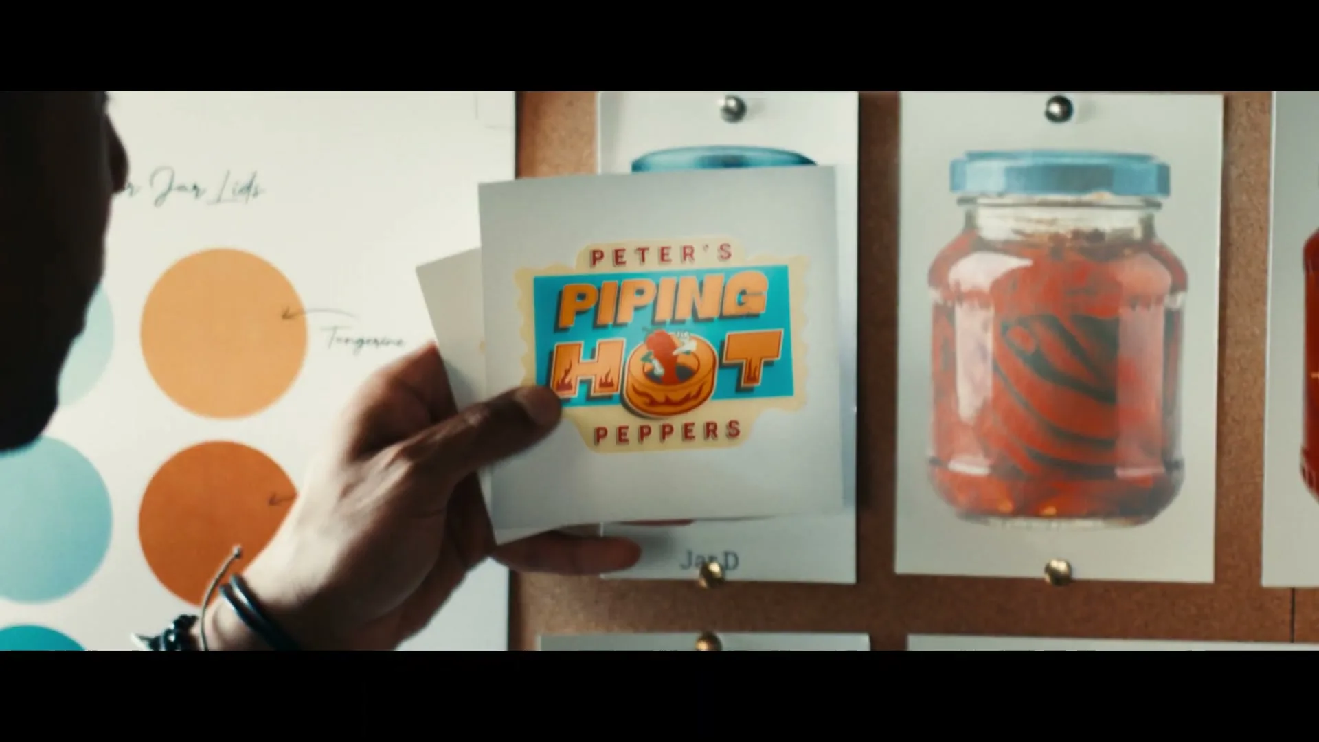 A man's hand holds up a "Peter's Piping Hot Peppers" label in front of a cork board with color swatches and jar designs, as he looks at it.