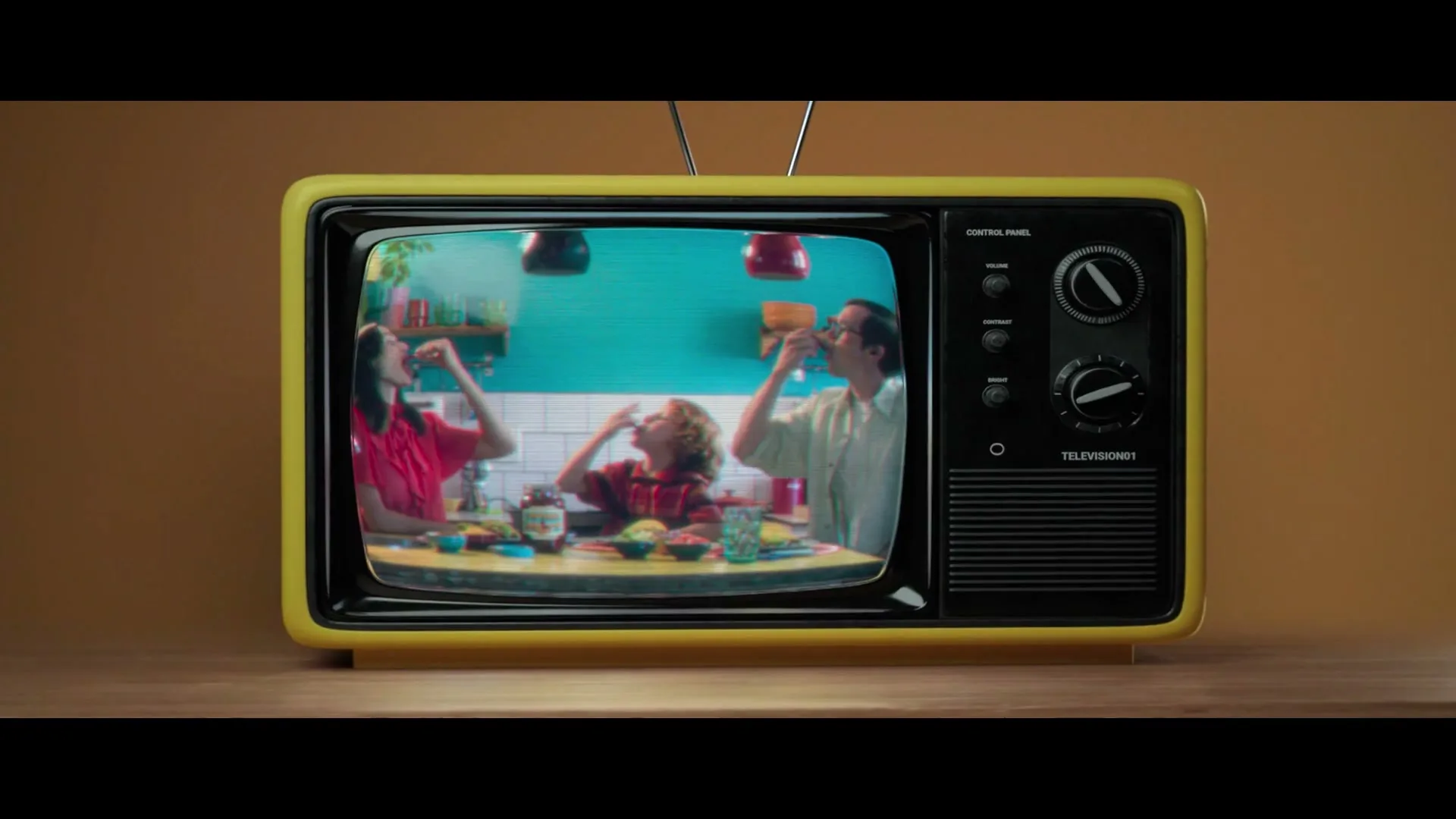 A retro yellow television set sits on a wooden surface, displaying a family eating around a table in a brightly lit kitchen.