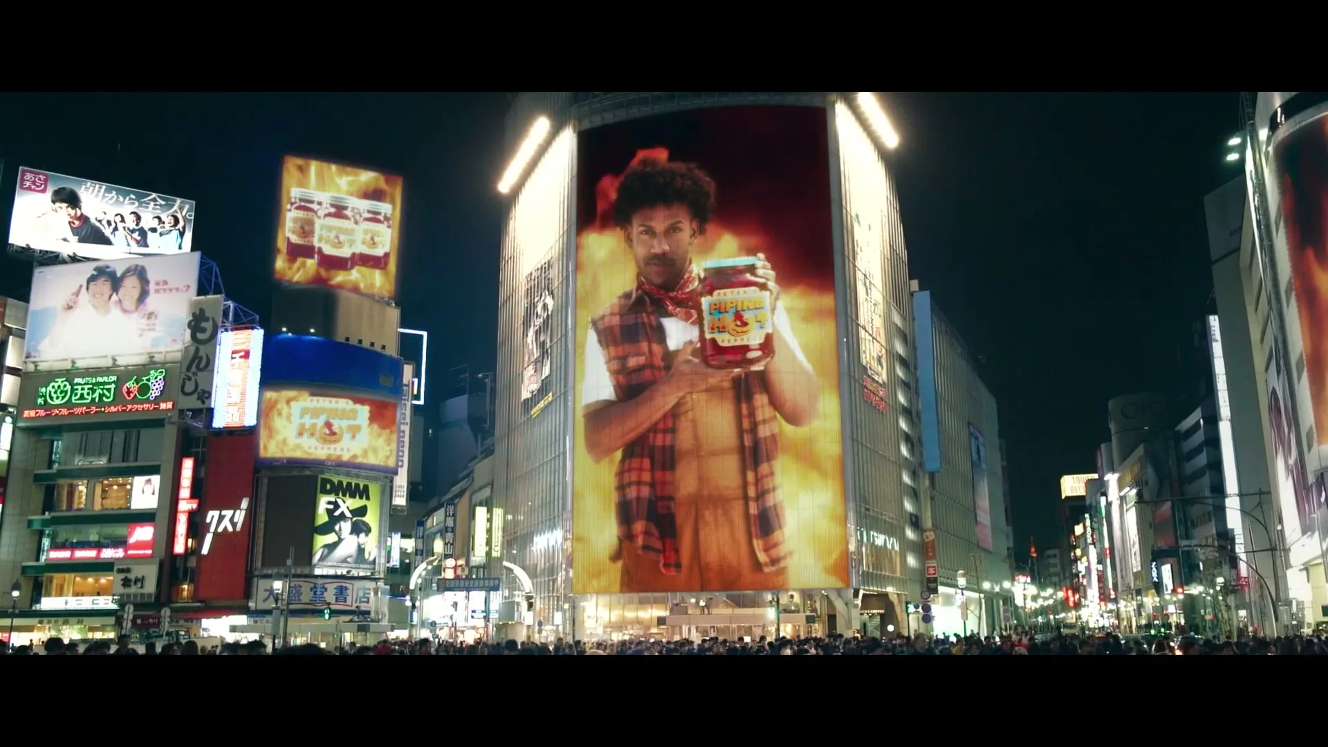 A man with curly hair and a plaid vest is shown on a giant billboard in a busy city square at night, holding a jar of pickles.