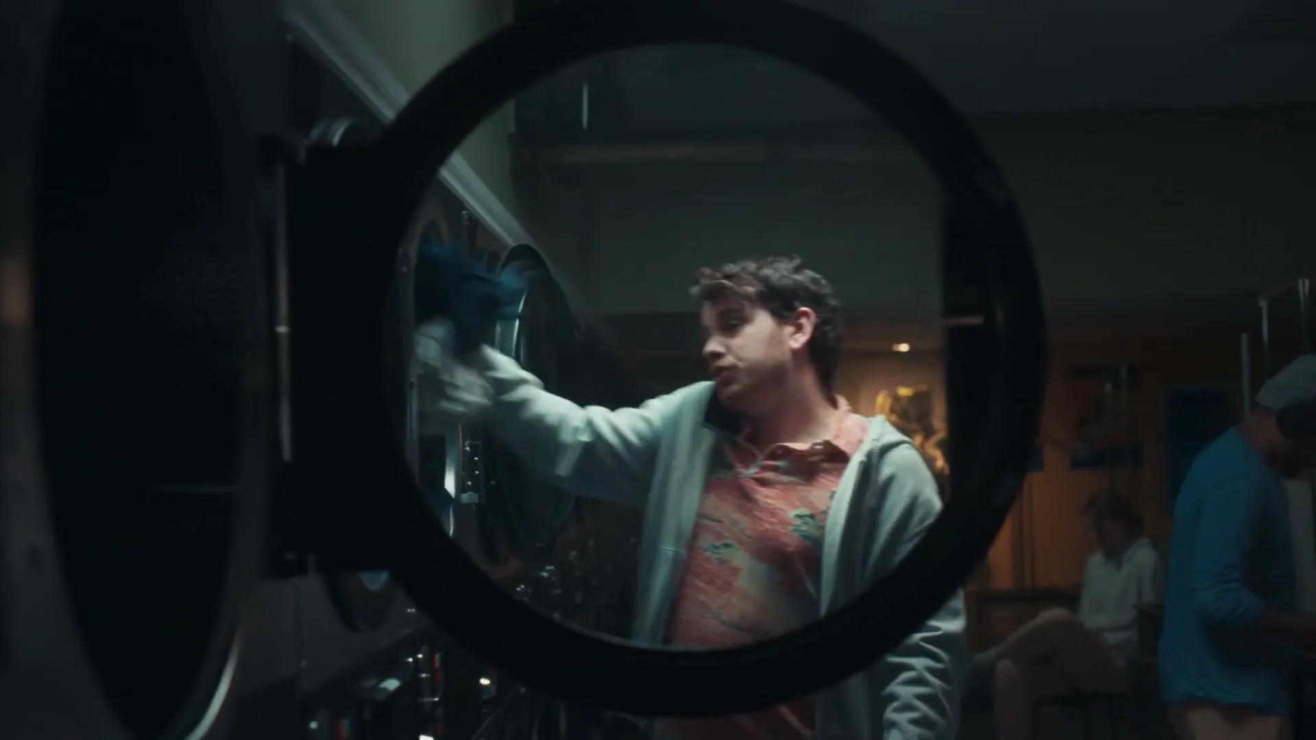 A young man in a grey hoodie and patterned shirt talks on the phone while doing laundry in a laundromat, seen through a washing machine door.