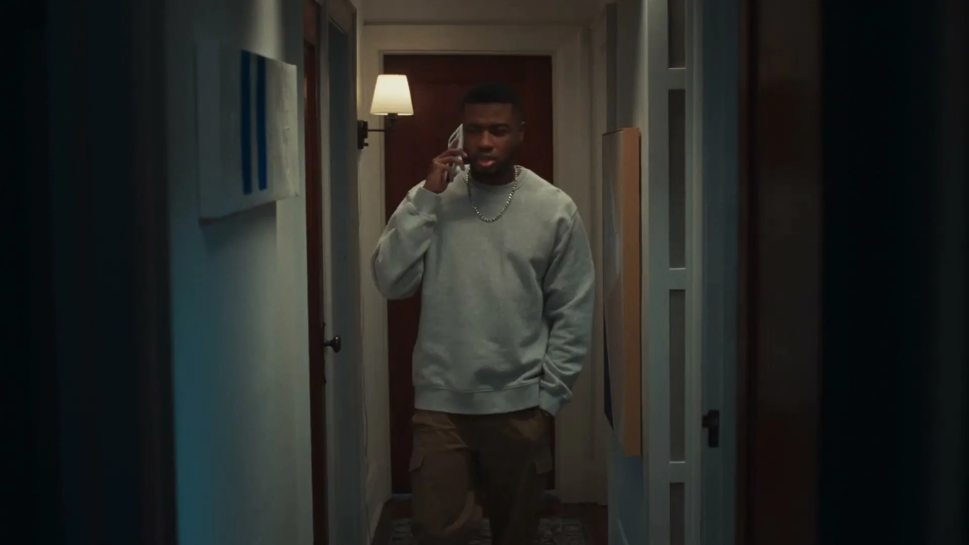 A young Black man in a grey sweatshirt and cargo pants talks on a white smartphone in a dimly lit hallway, looking confused.