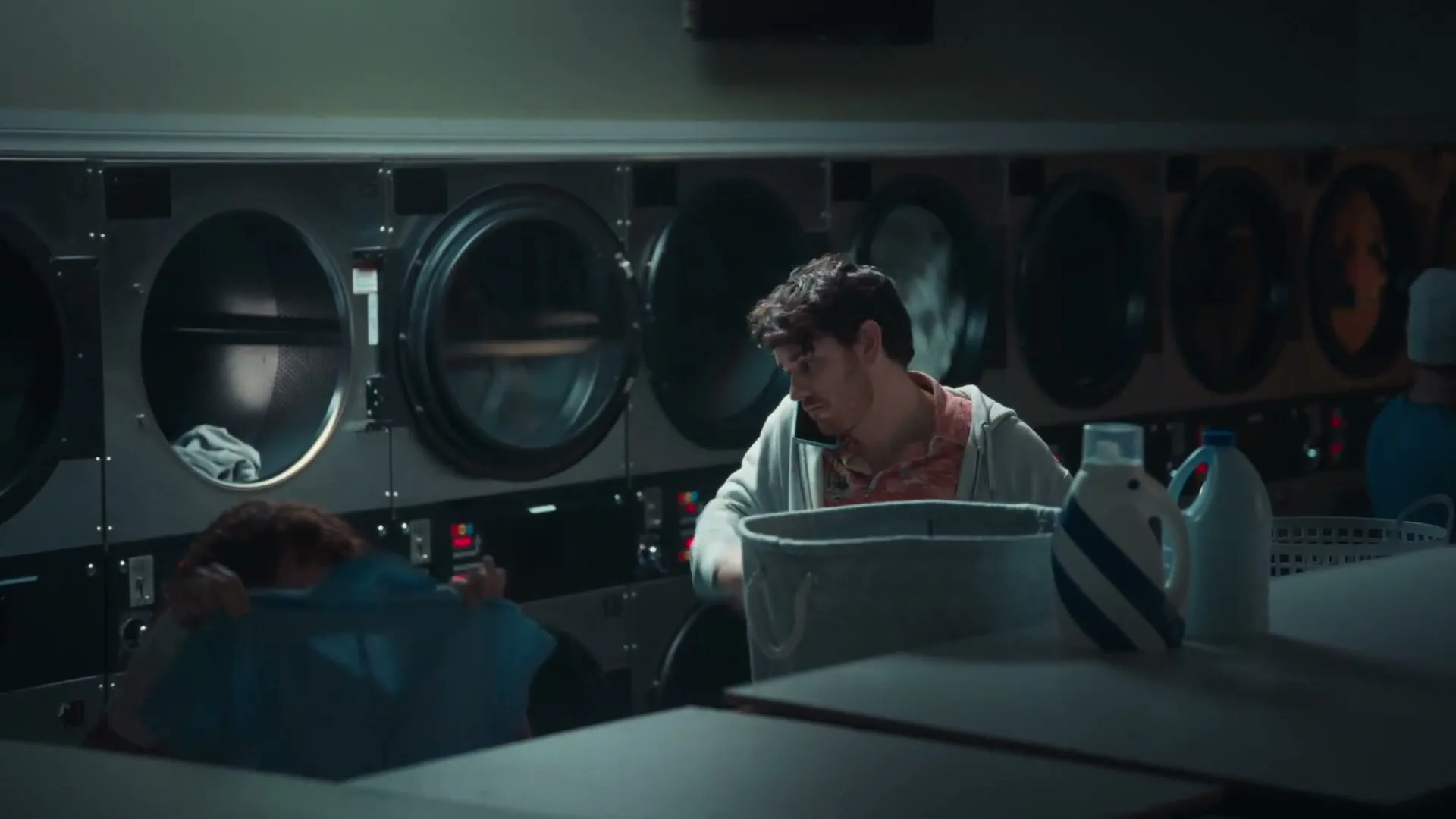 A man talks on his phone while doing laundry in a laundromat, as a young girl sits nearby, looking down.