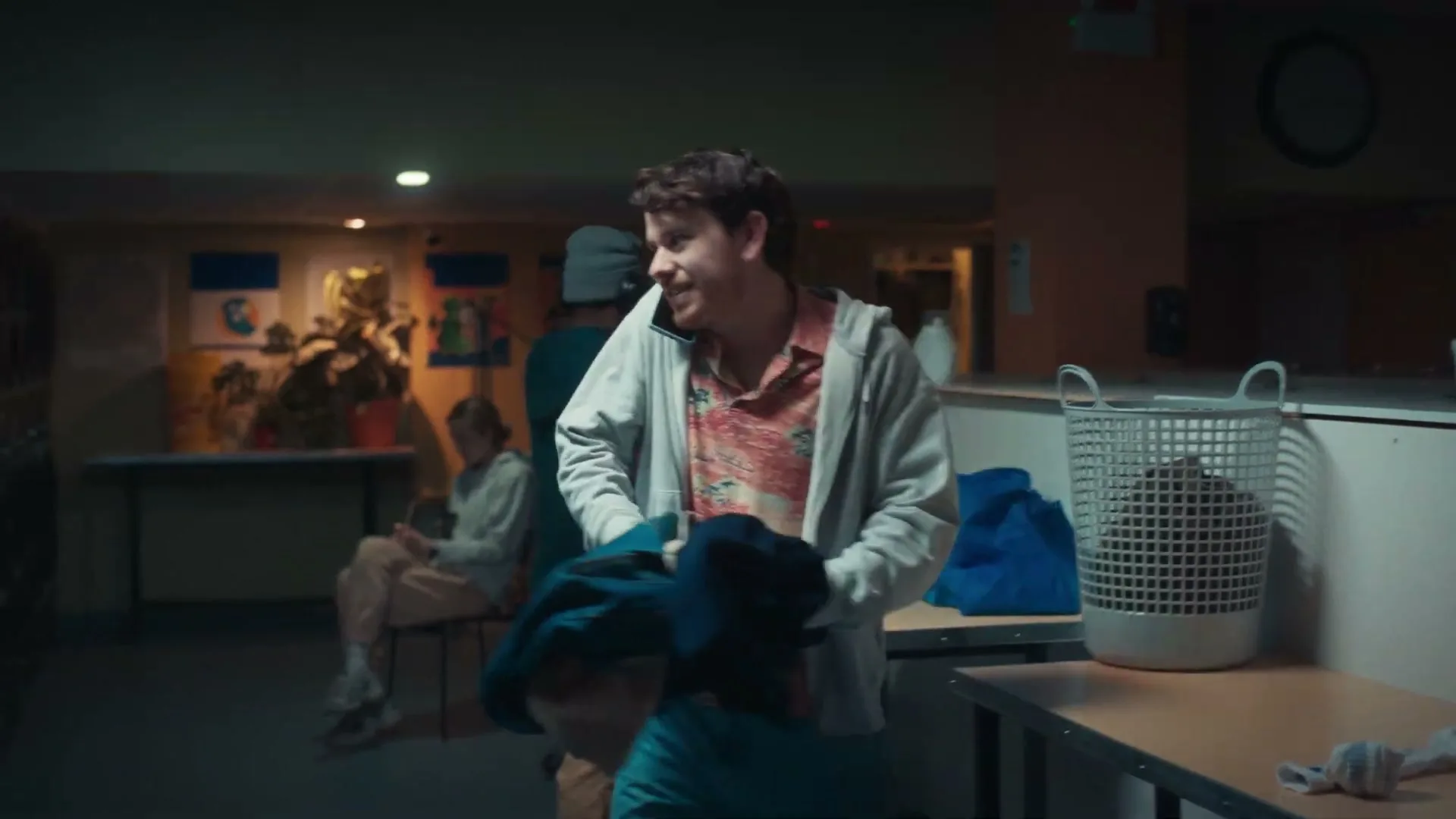 A young man in a hoodie talks on the phone while doing laundry in a dimly lit laundromat, looking around with a confused expression.