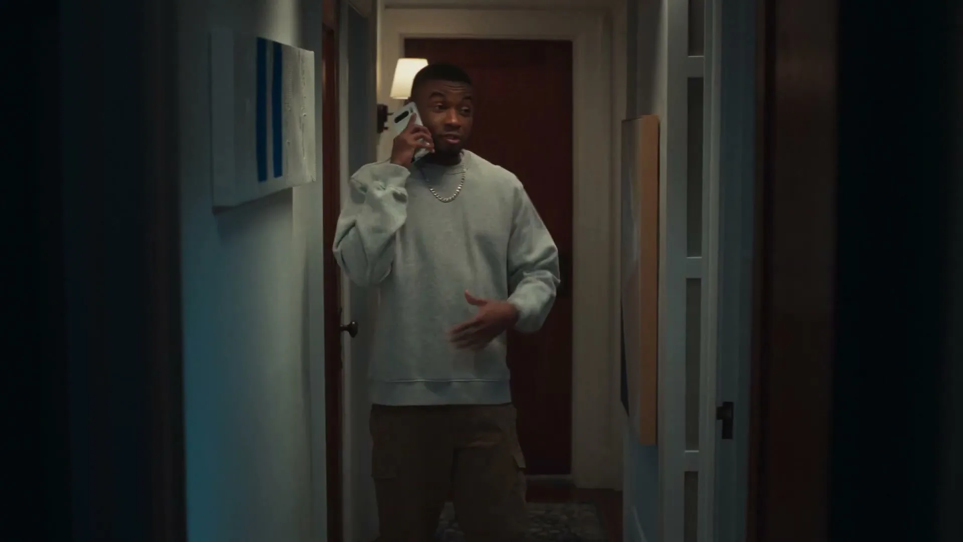 A young Black man in a grey sweatshirt and cargo pants talks on a white smartphone in a dimly lit hallway, gesturing with his free hand.