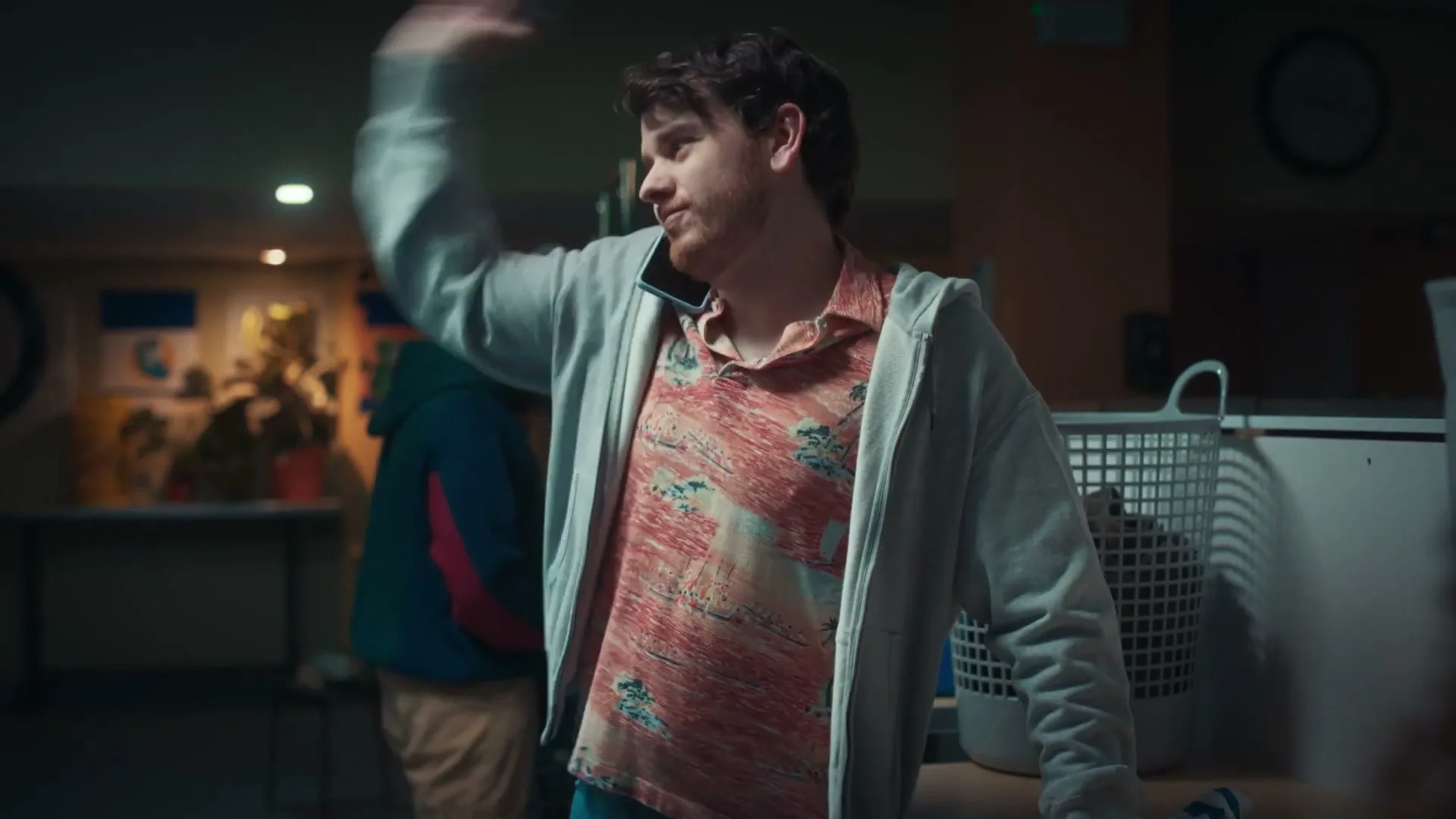 A young man in a Hawaiian shirt and hoodie talks on a phone, looking frustrated, in a laundromat at night.