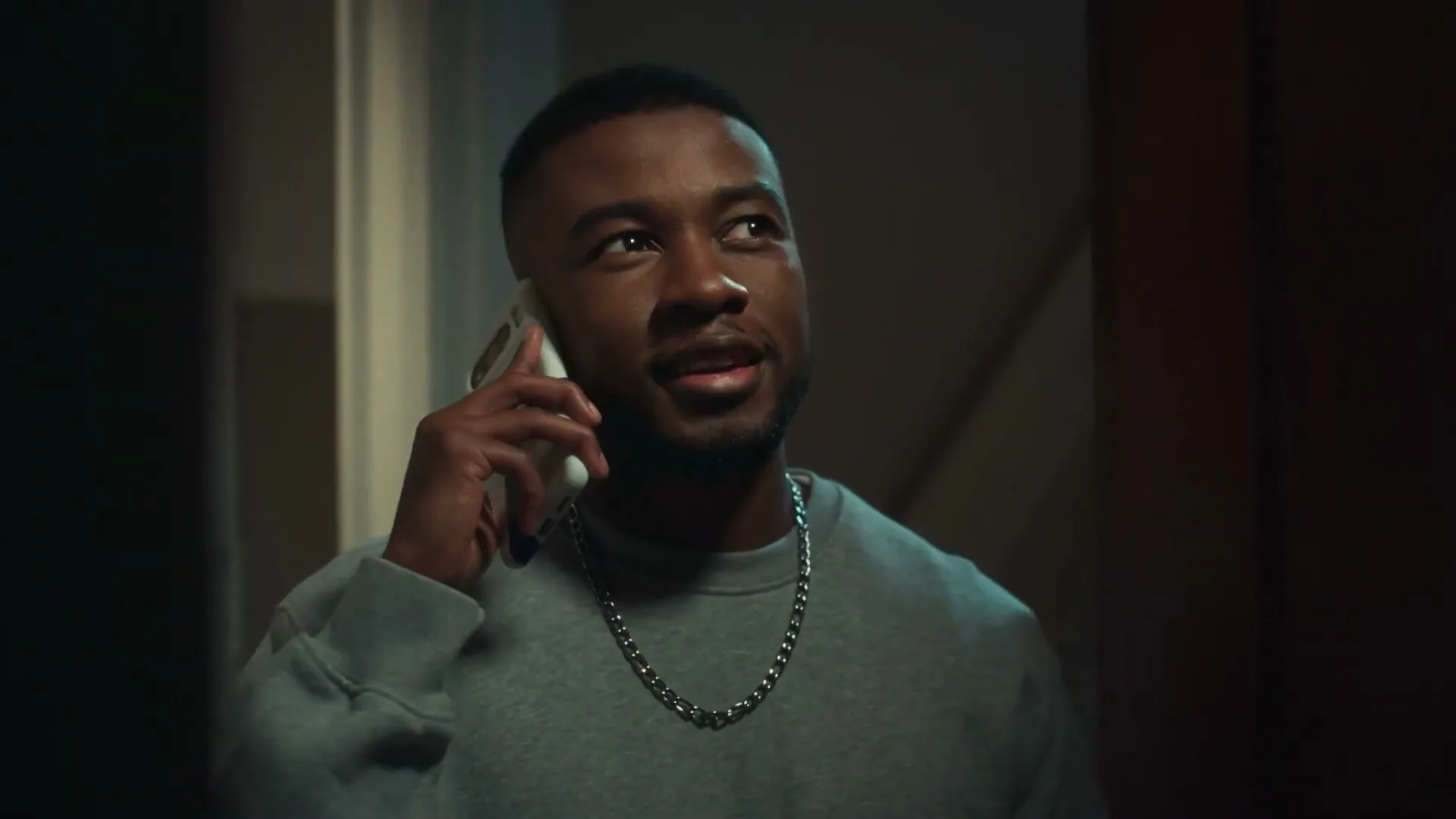 A young Black man in a grey sweatshirt and silver chain talks on a white smartphone, looking to the right in a dimly lit interior.
