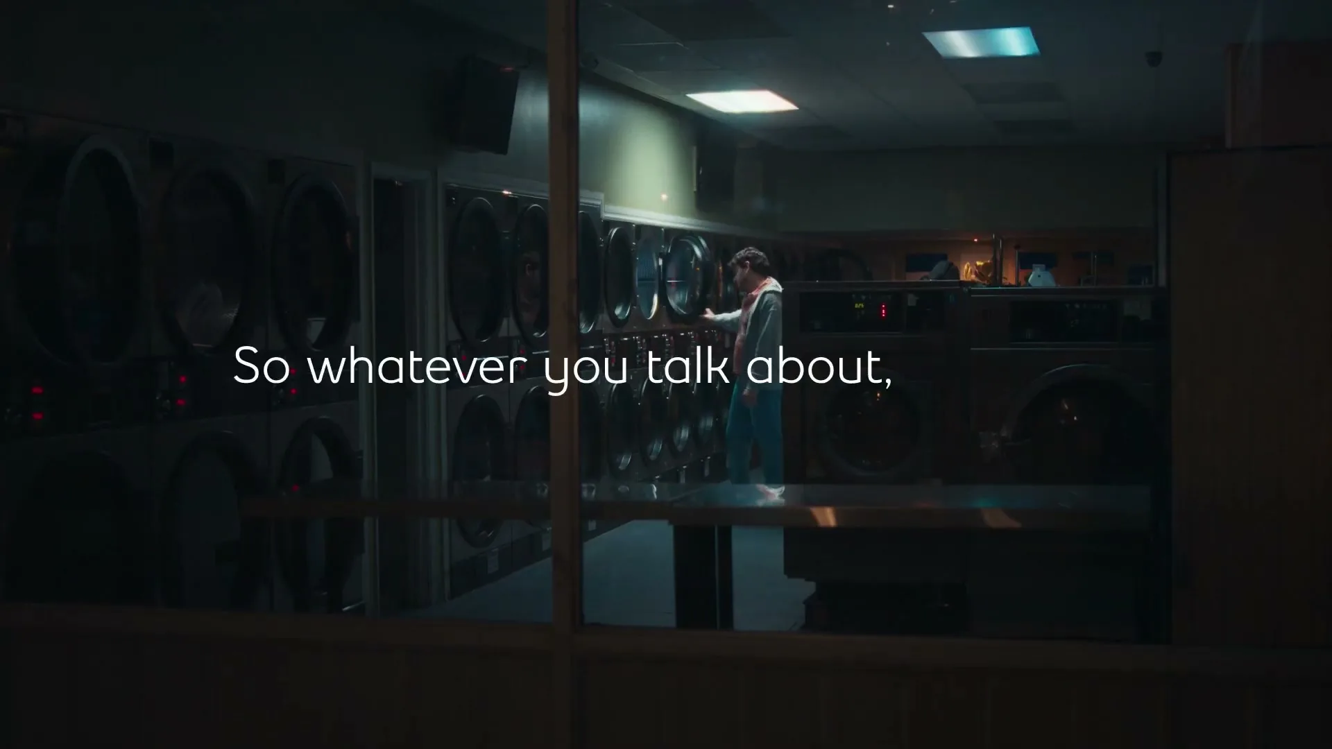 A young man in a hoodie and jeans stands in a laundromat, talking on the phone, seen through a window at night.