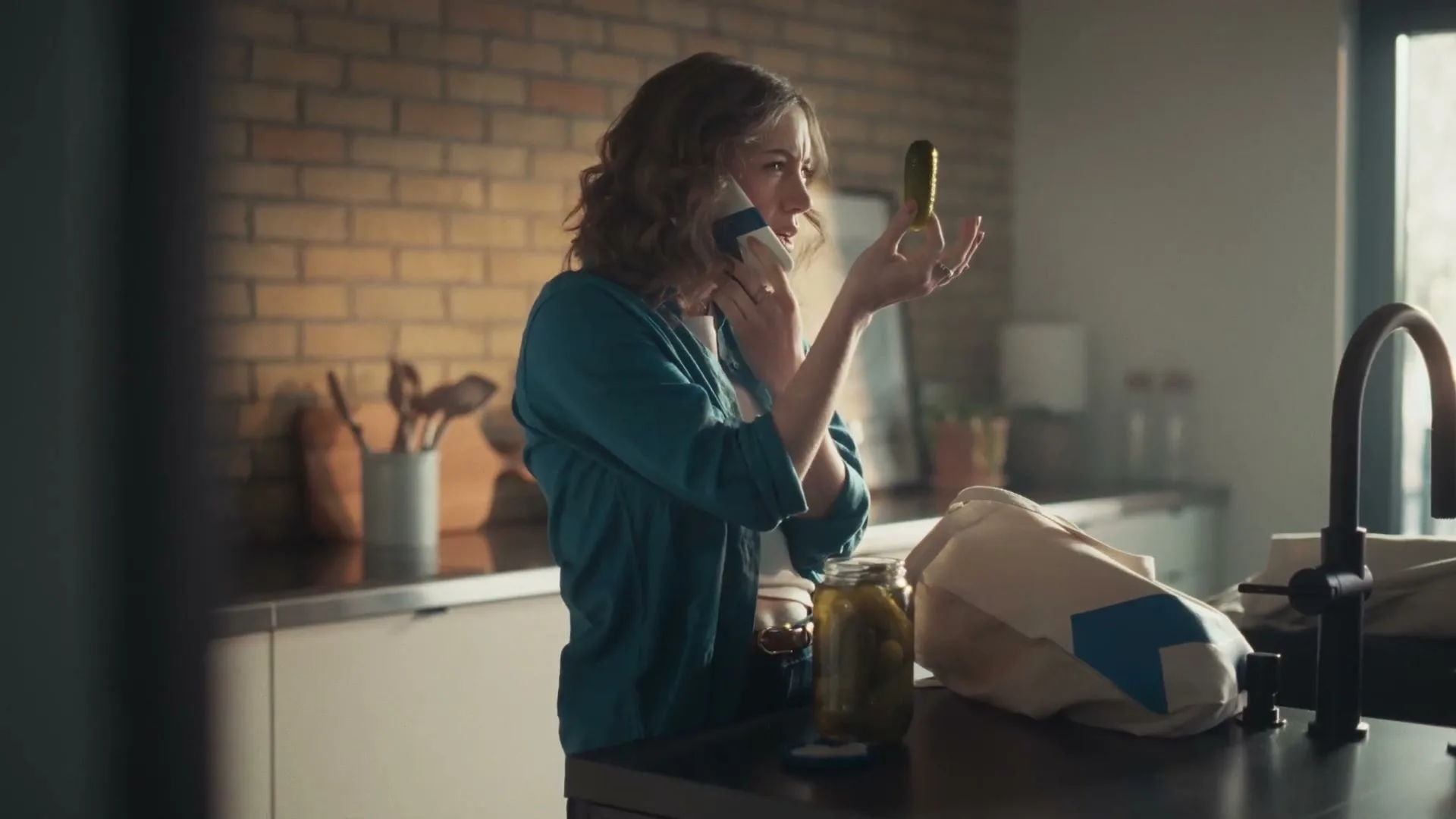 A woman in her 30s, wearing a teal shirt, talks on the phone while holding a pickle and looking confused in a modern kitchen.