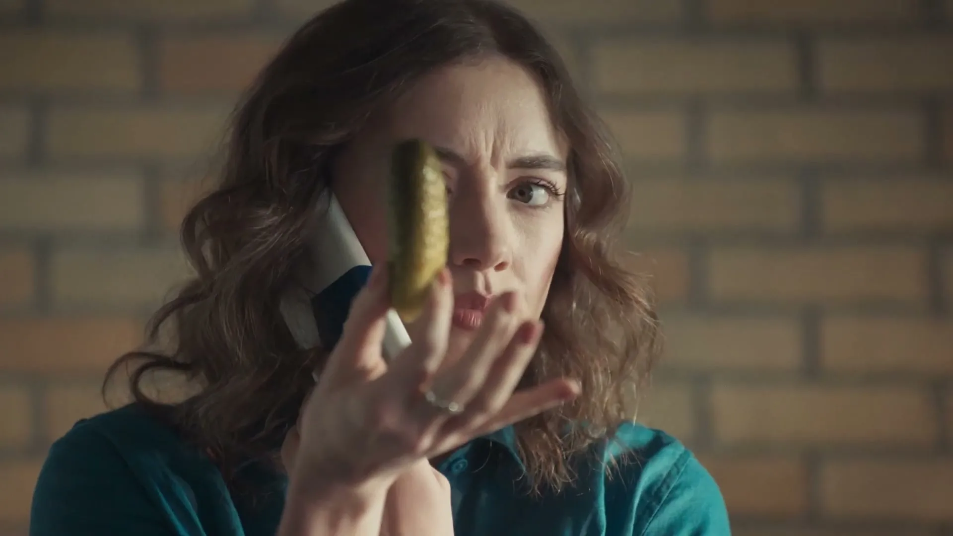 A woman with brown hair holds a pickle in front of her face while talking on the phone, looking confused in an interior setting.