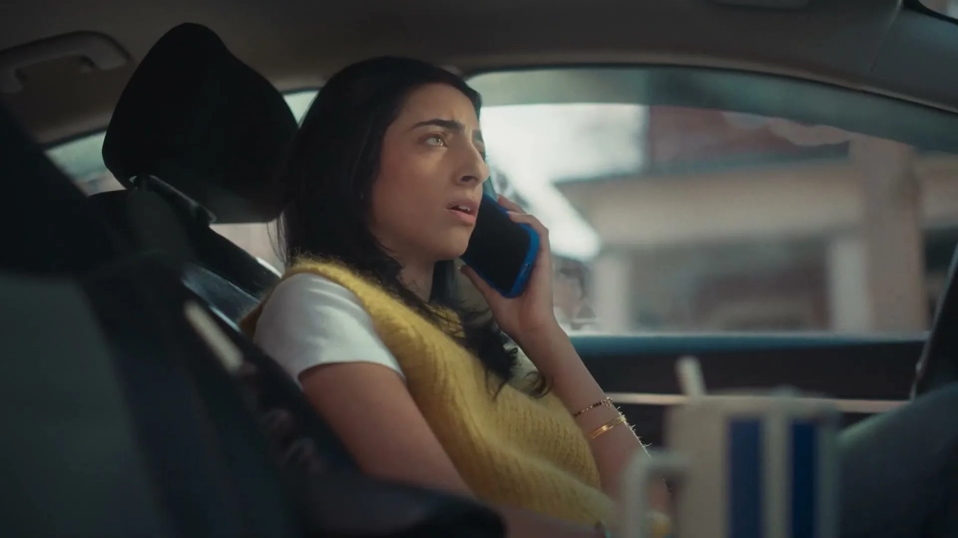 A young woman with dark hair, wearing a yellow vest over a white t-shirt, sits in a car and talks on a blue phone, looking concerned.