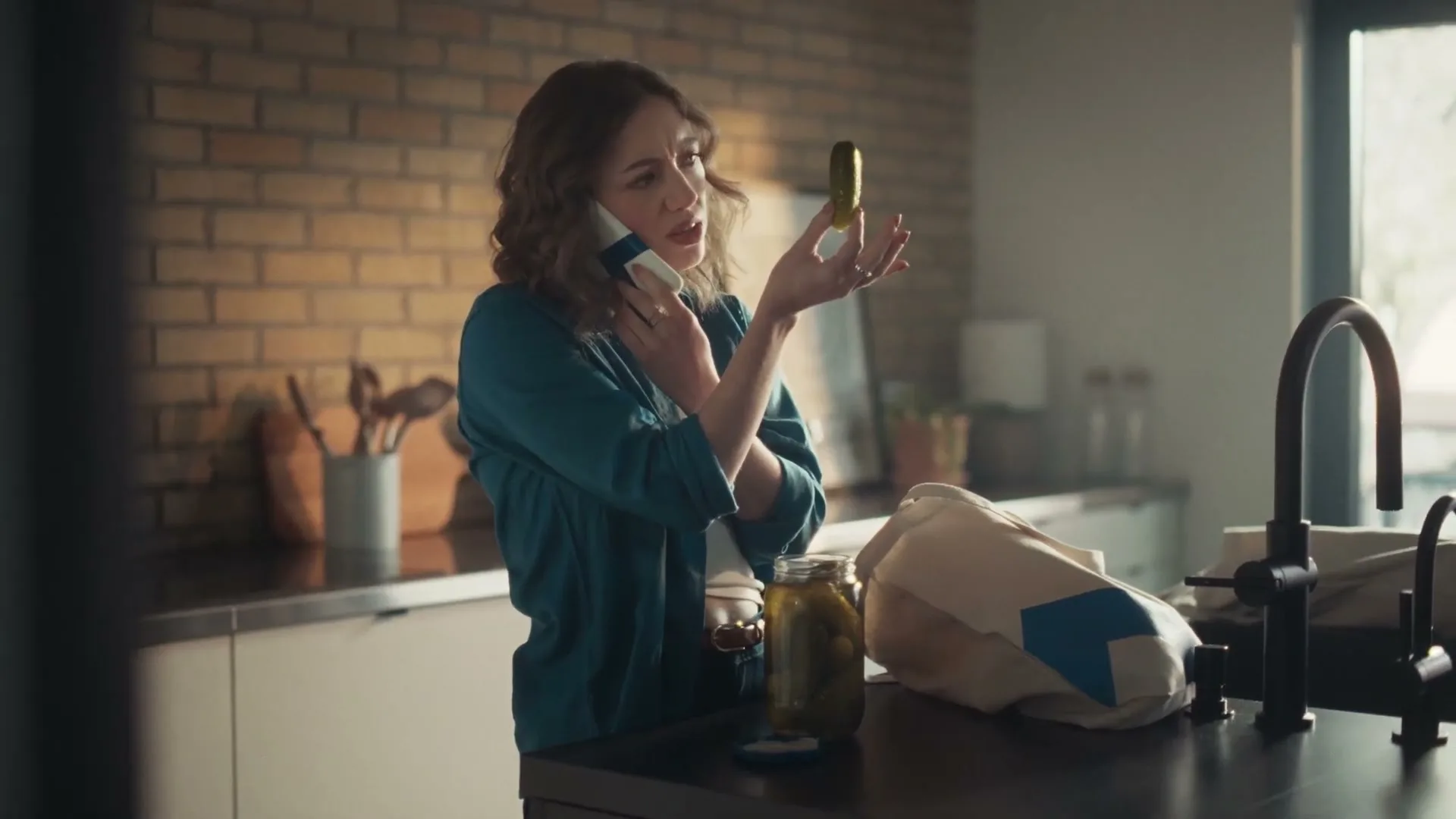 A woman in her 30s, wearing a teal shirt, talks on a phone while holding a pickle in a modern kitchen.
