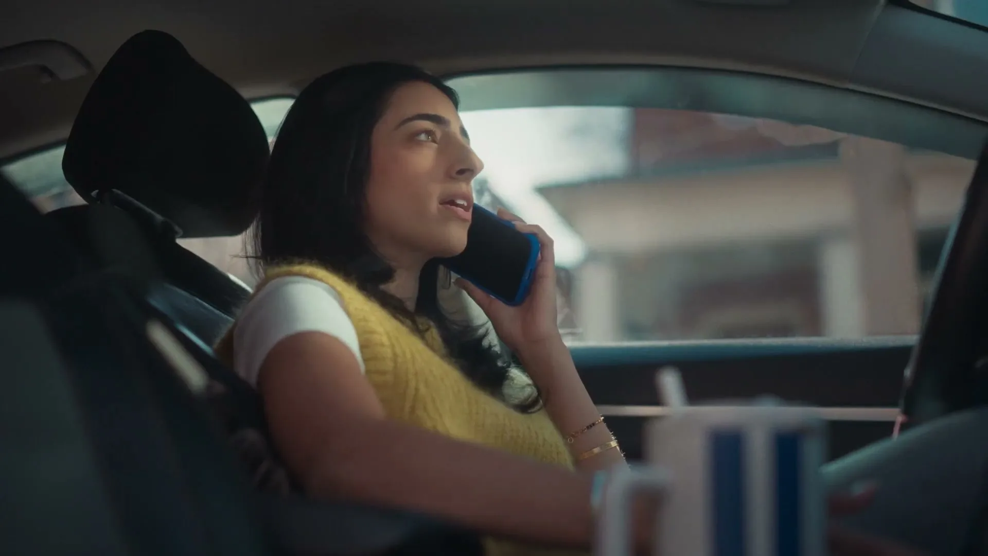 A young woman with dark hair, wearing a yellow sweater, sits in a car and talks on a phone, looking thoughtful.