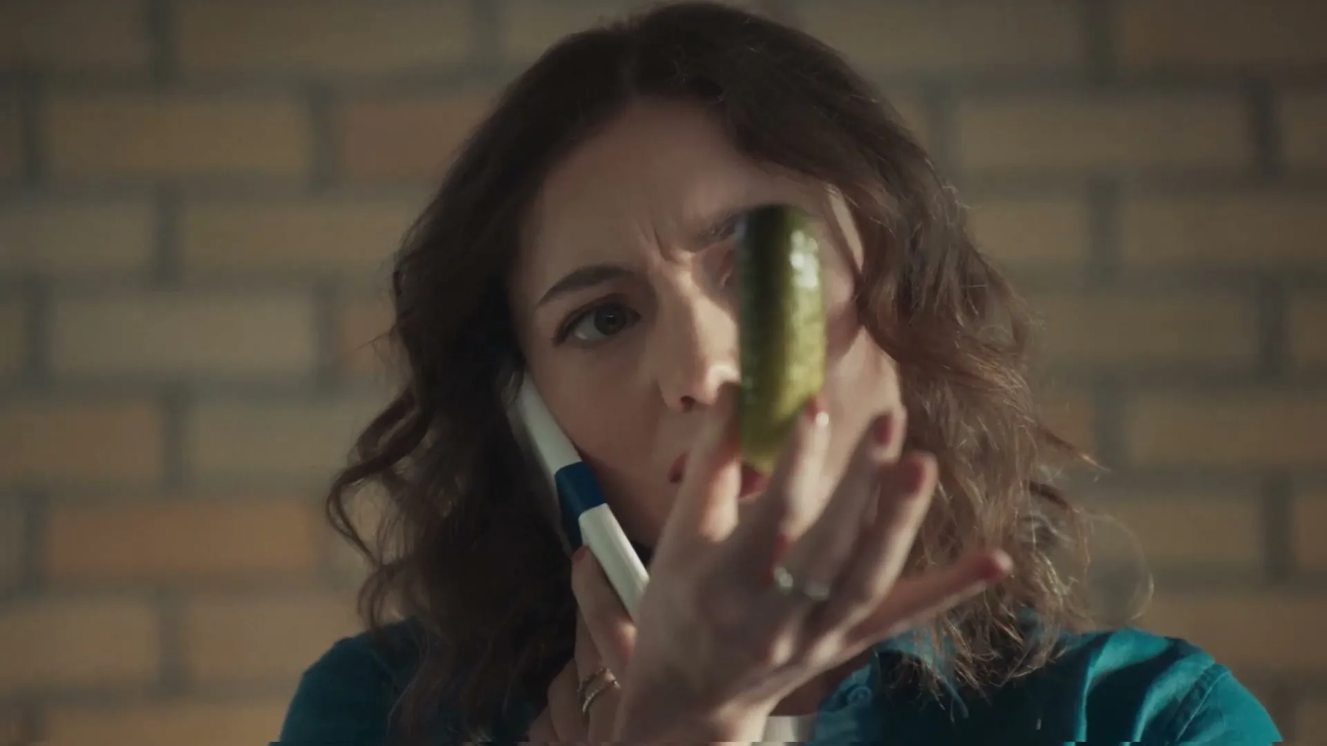 A young woman with curly brown hair holds a phone to her ear and a pickle in front of her face, looking confused in an interior setting.