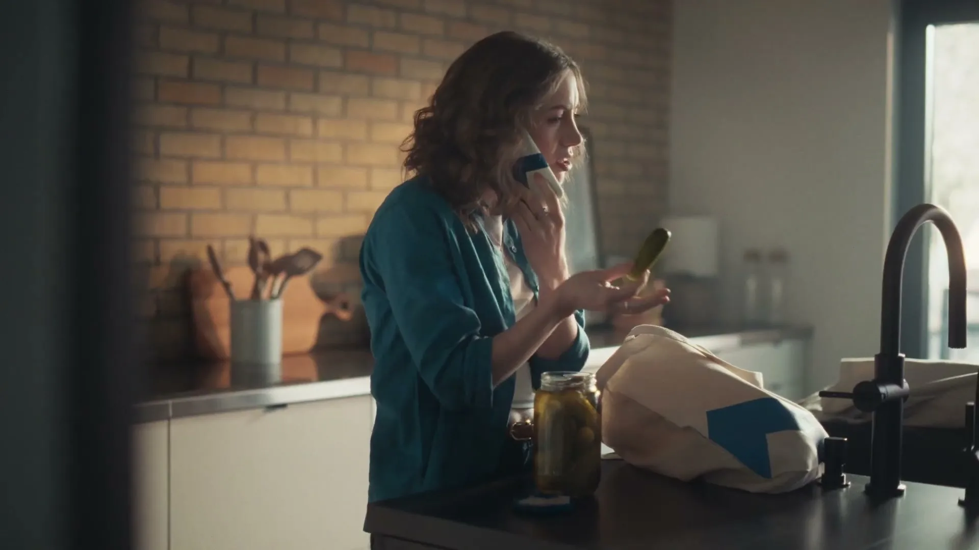 A woman in her 30s, wearing a teal shirt, talks on the phone while holding a pickle in a modern kitchen.
