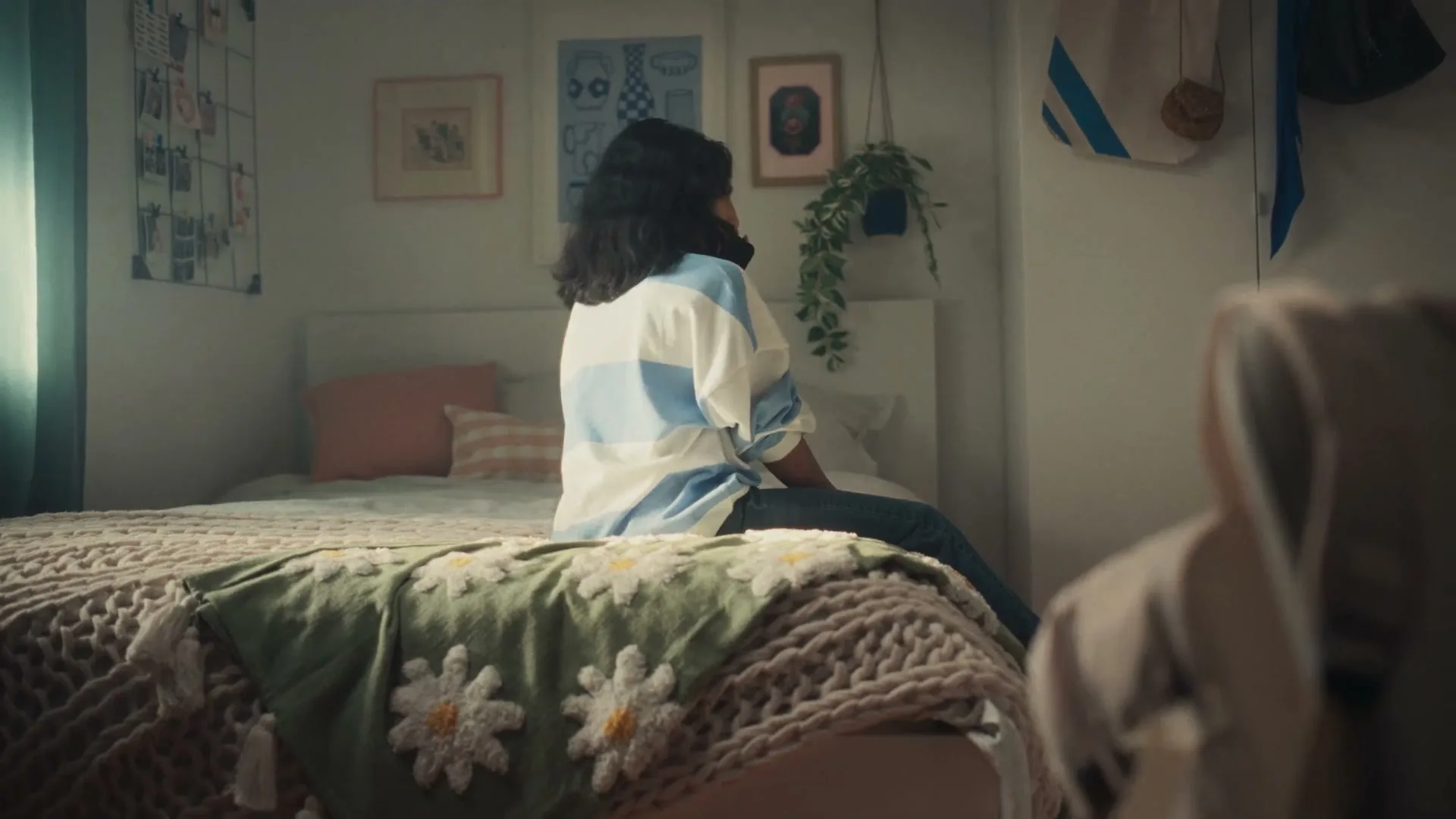 A young woman with dark hair sits on a bed in a cozy bedroom, talking on a phone, with a crocheted blanket in the foreground.