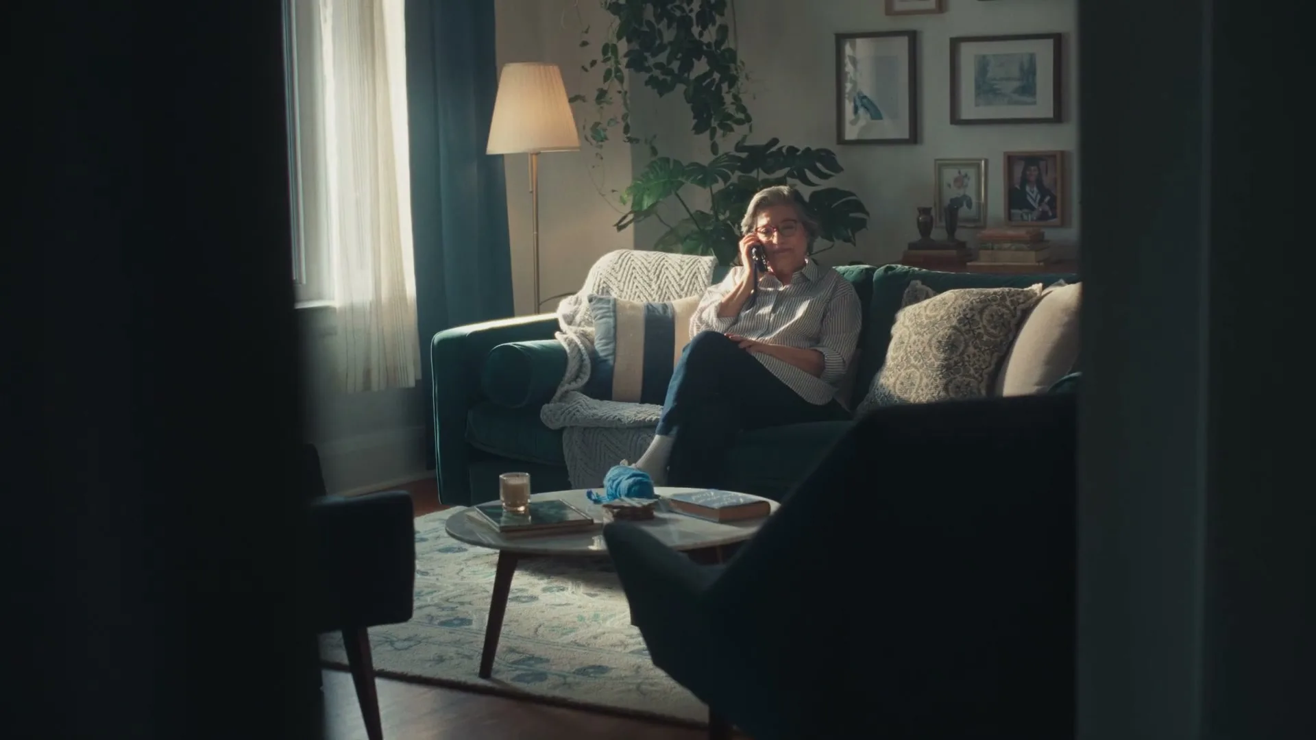 A senior woman in a striped shirt and jeans sits on a couch, talking on a phone in a cozy living room.