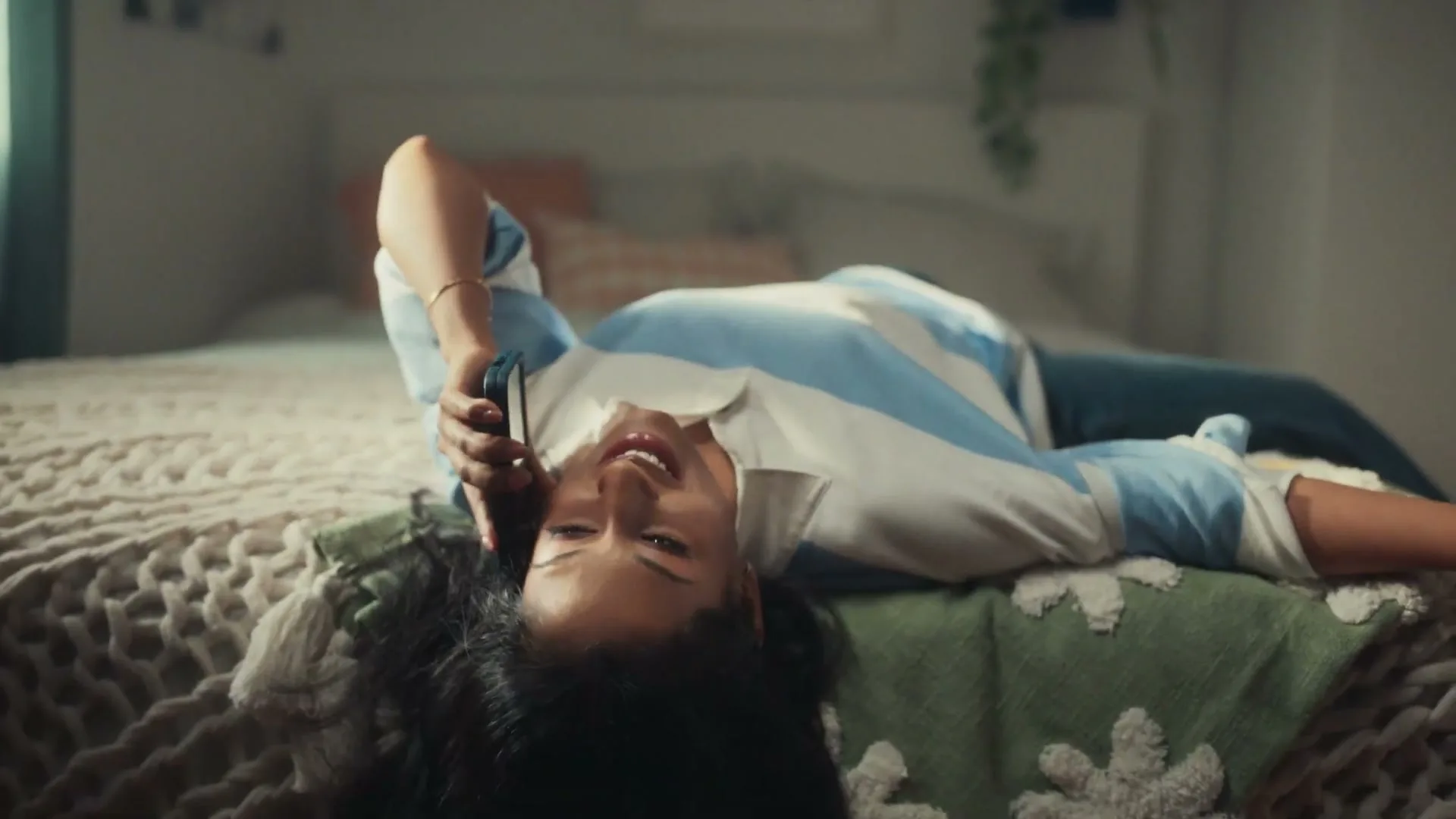 A young woman with dark hair, wearing a striped shirt, lies on her bed, talking on her phone and smiling in her bedroom.