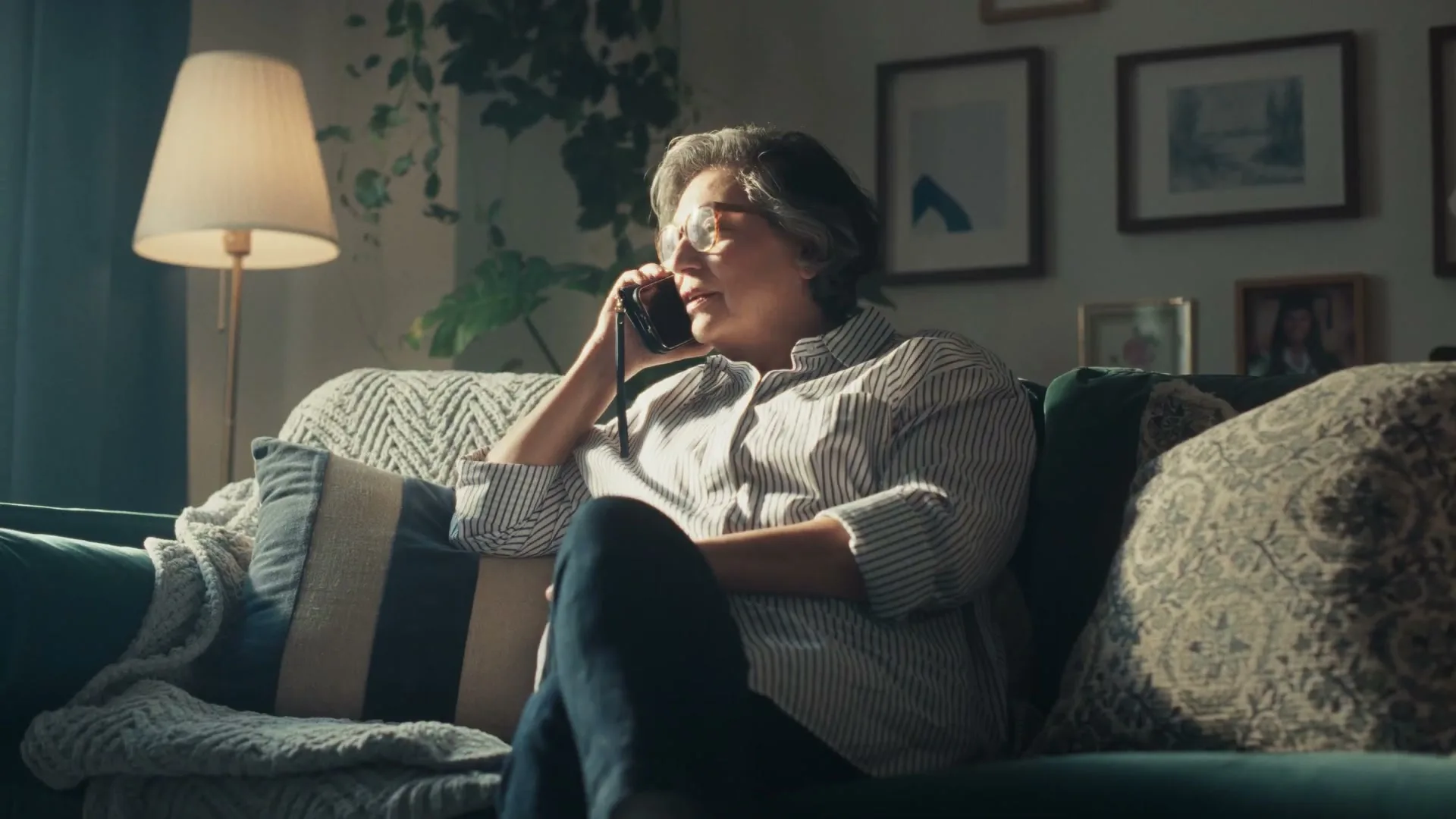 A woman in her 60s sits on a couch, talking on the phone, looking up and to the right, in a sunlit living room.