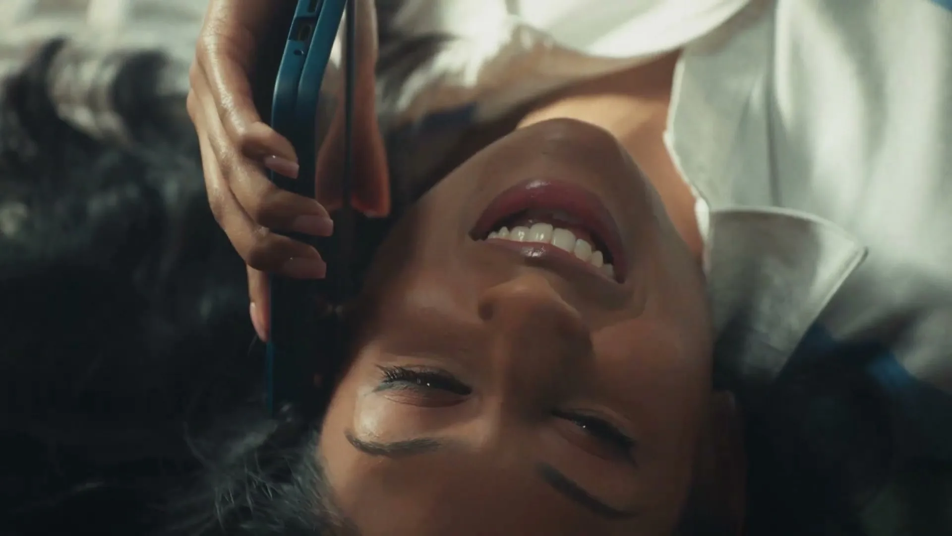 A young woman with dark hair and a white shirt lies down, holding a blue phone to her ear, smiling with her eyes closed.