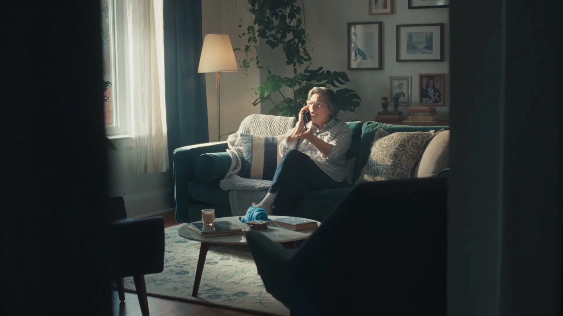 A woman in her 50s sits on a teal couch in a sunlit living room, talking on a phone.