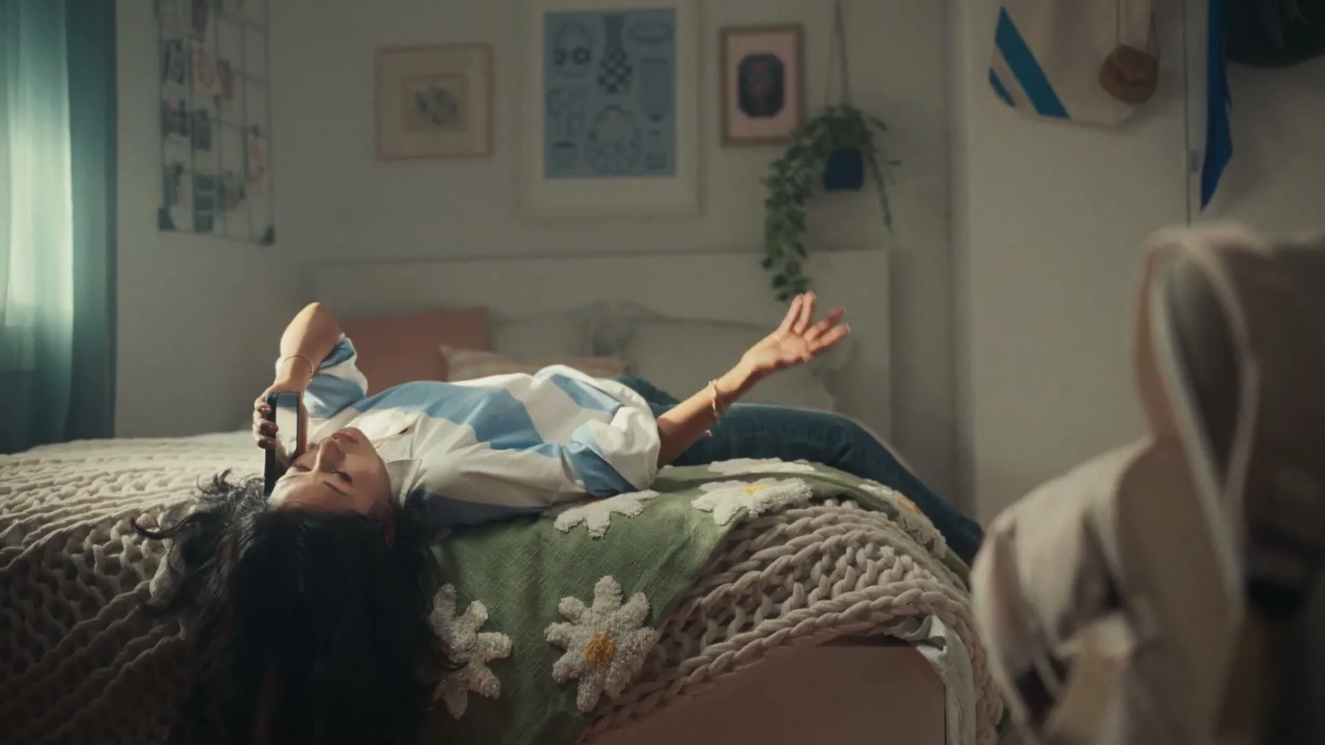 A young woman with long dark hair lies on a bed, talking on her phone and gesturing with her hand in a cozy bedroom.