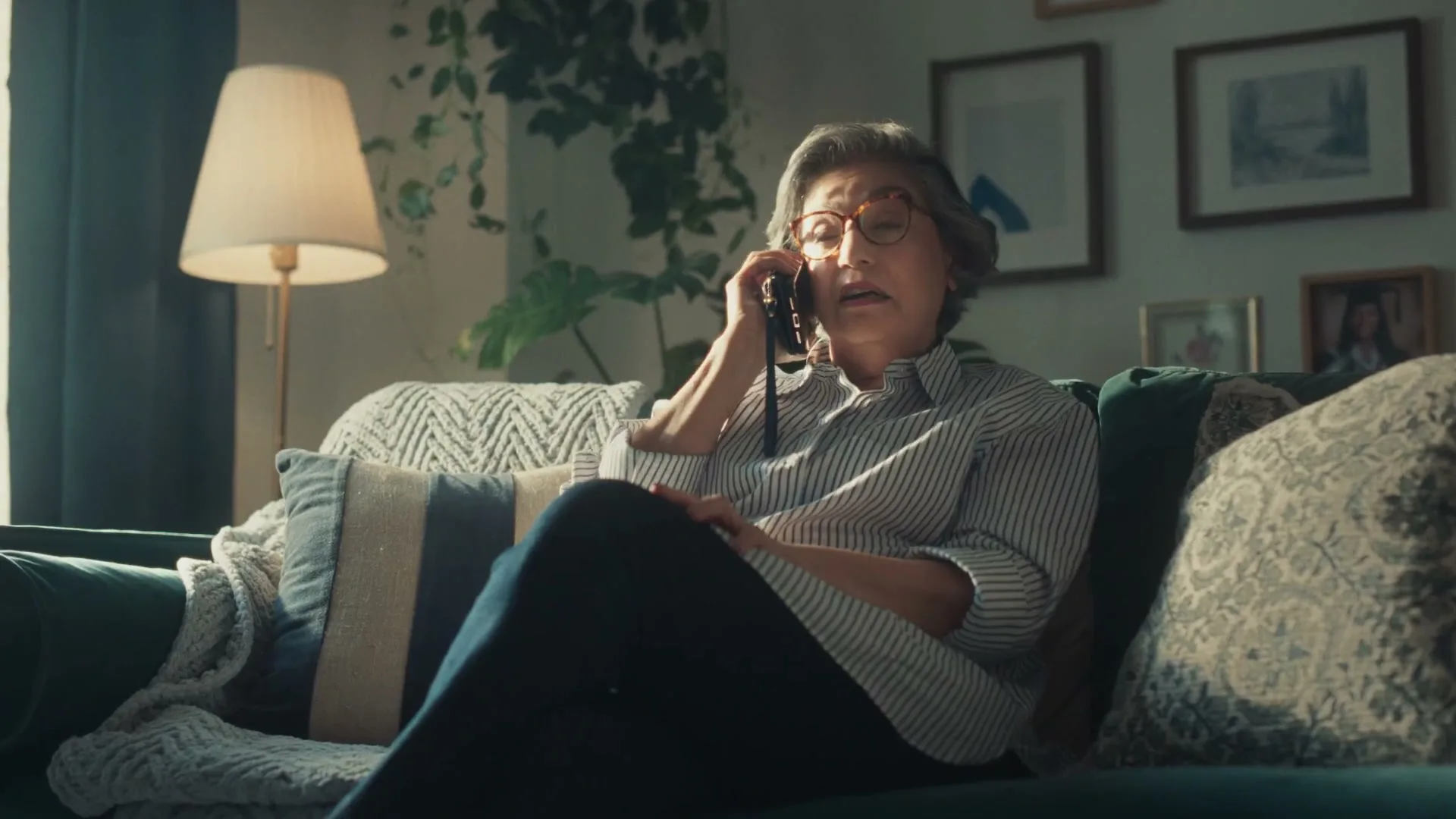 A woman in her 60s sits on a green couch, talking on a phone with a surprised expression in a cozy living room.