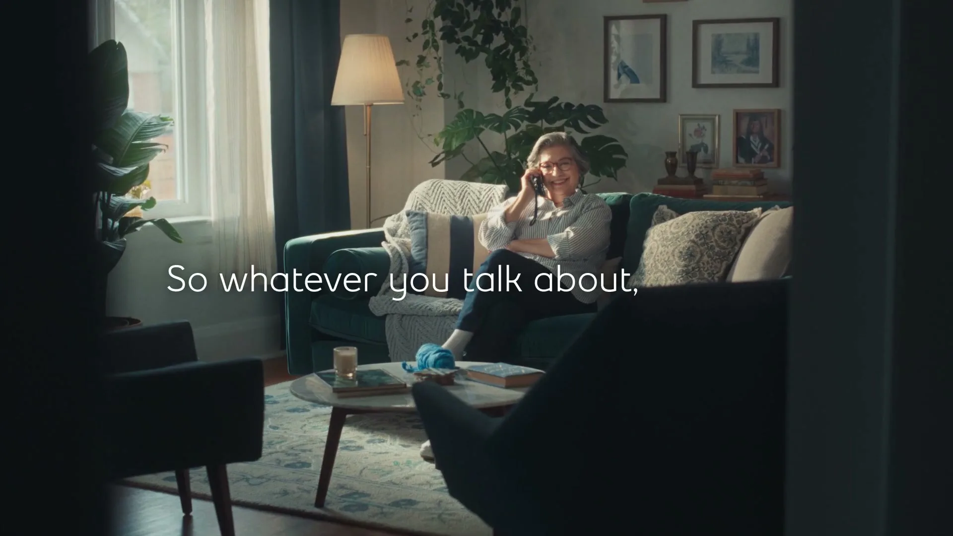A senior woman with glasses and a striped shirt sits on a teal couch, talking on a phone and smiling, in a cozy living room.