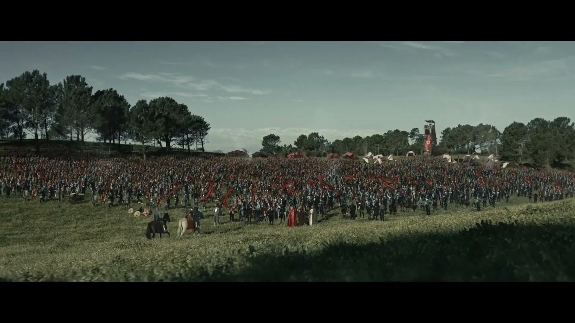 A vast army of soldiers in historical attire stands on a grassy battlefield under a cloudy sky, with tents and a tower in the background.