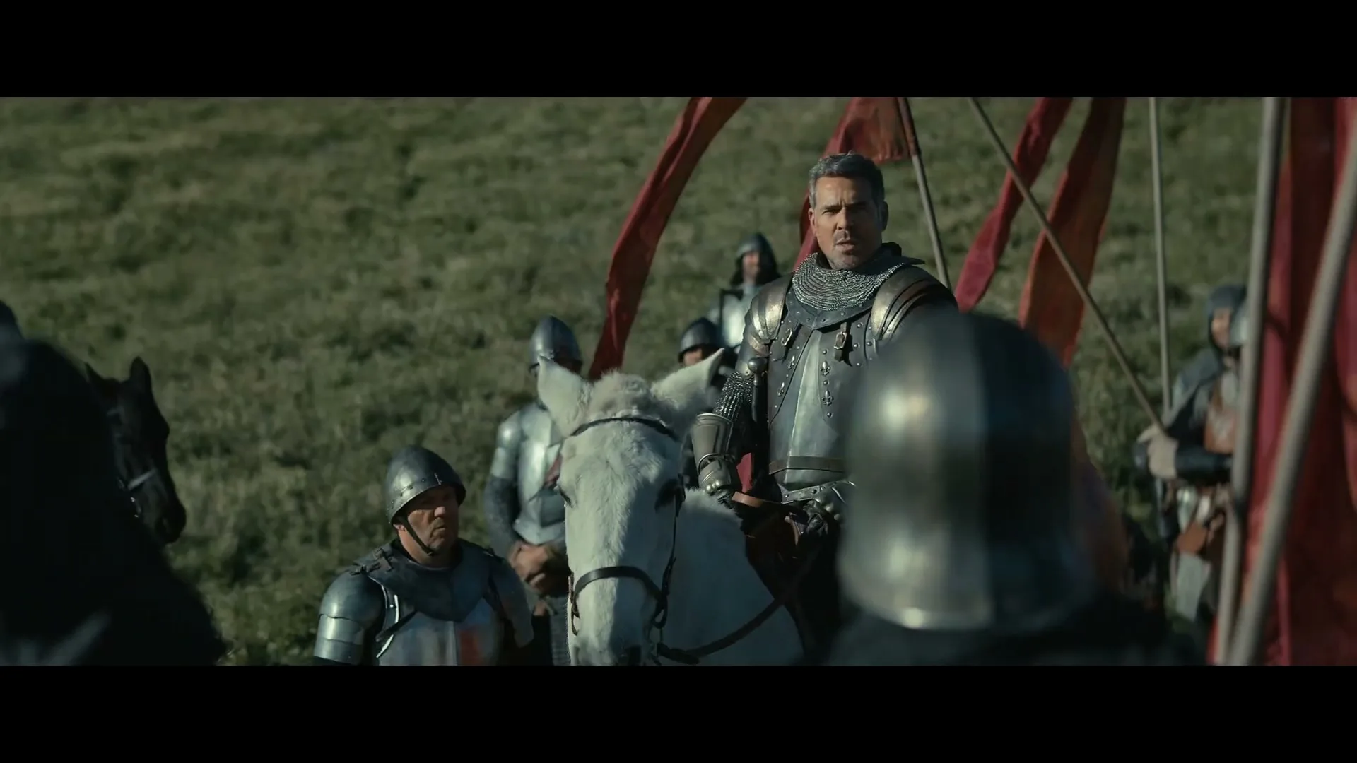 A male knight in shining armor on a white horse addresses his troops on a grassy battlefield under a bright sky.